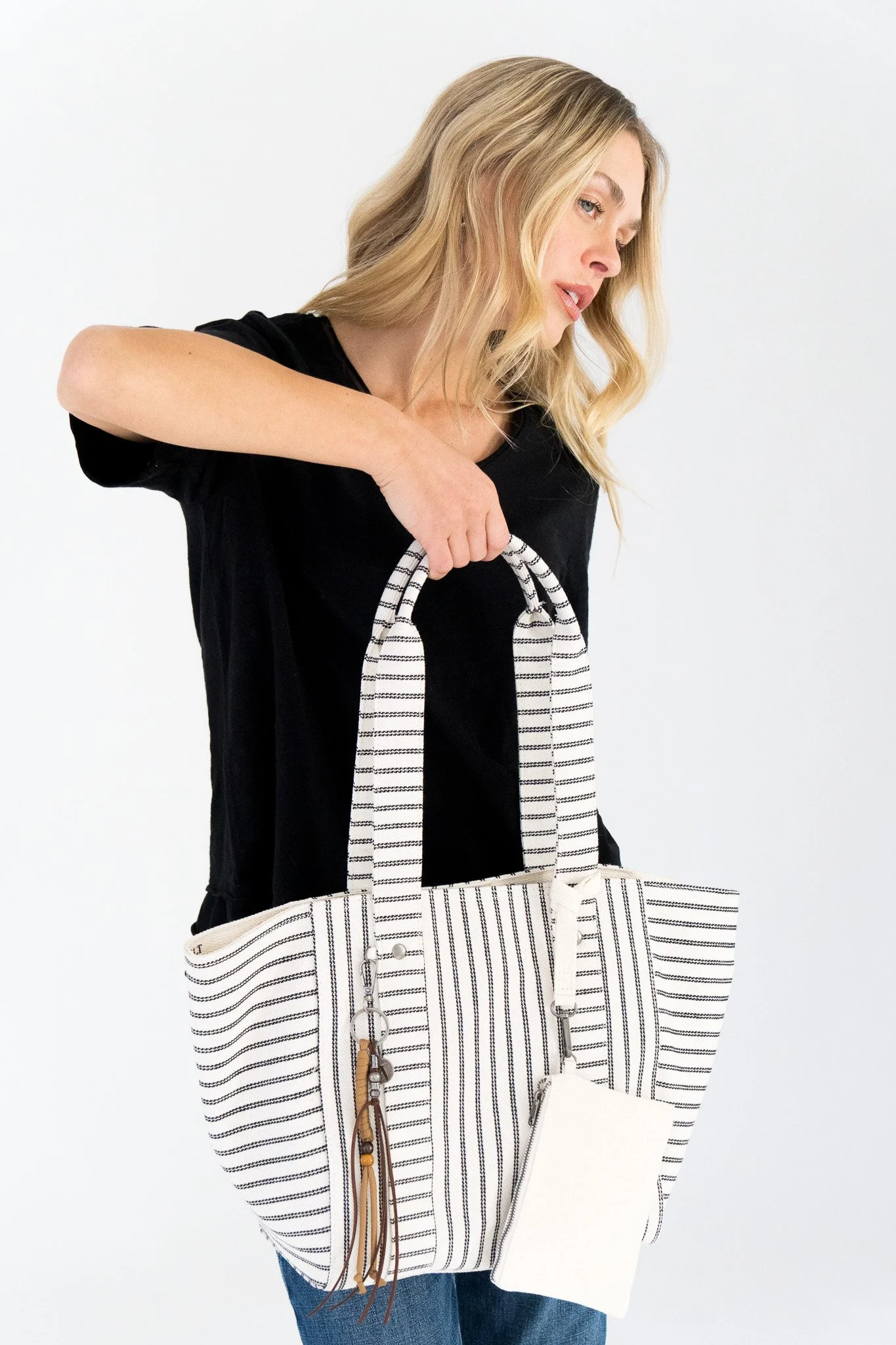 Woman wearing a black top, holding a striped bag with leather and beads, and a white bag, against a plain white background.