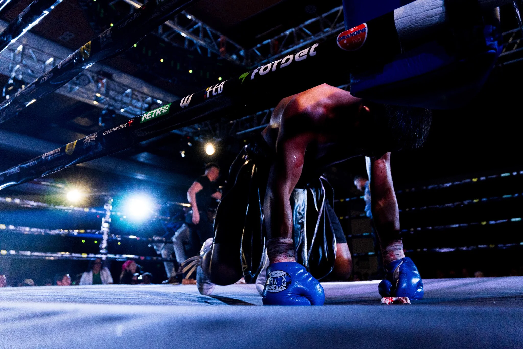 A boxer kneeling in the ring with blood on his face, wearing blue gloves and shorts, under bright lights with a dark background, and a person in the background.