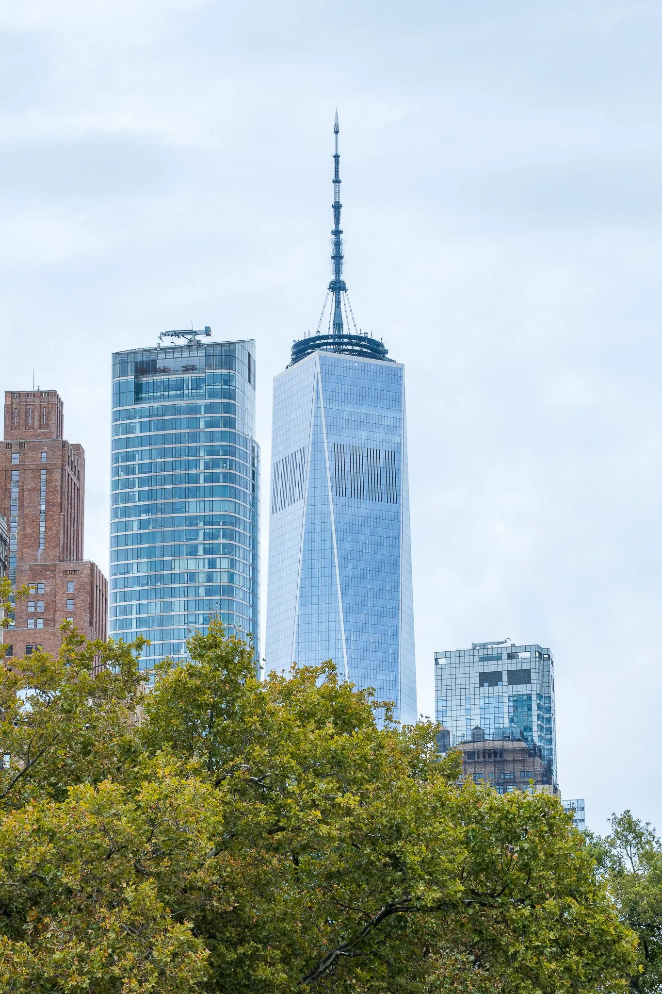 Skyline of NYC with One World Trade Center and surrounding buildings, green trees in foreground.