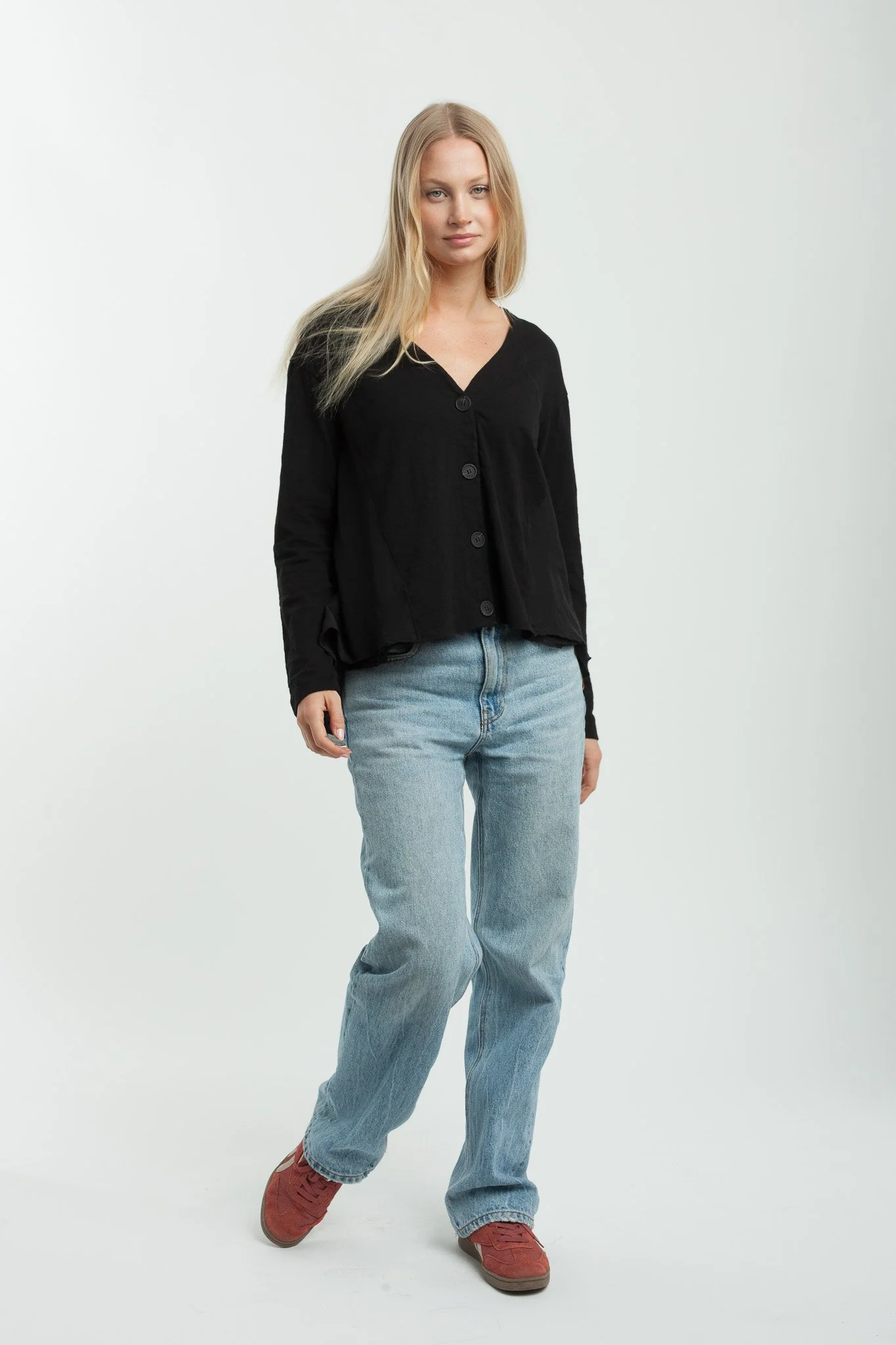 A young woman with long blonde hair wearing a black button-up shirt, light blue jeans, and brown shoes standing against a plain white background.