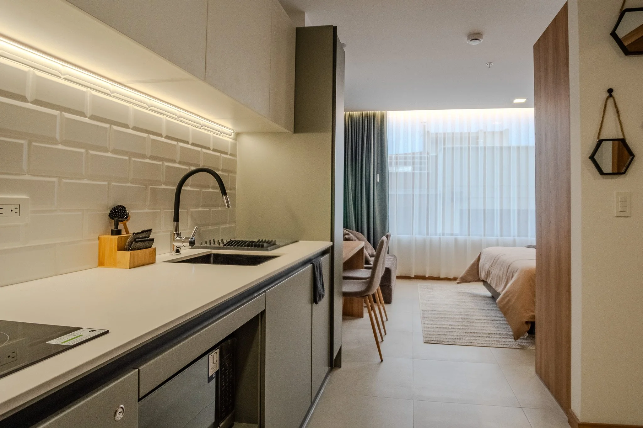 A modern kitchen with white countertop, black sink, wooden knife block, and beige cabinets, leading into a living space with a bed, window with sheer curtains, and a dining area with chairs.