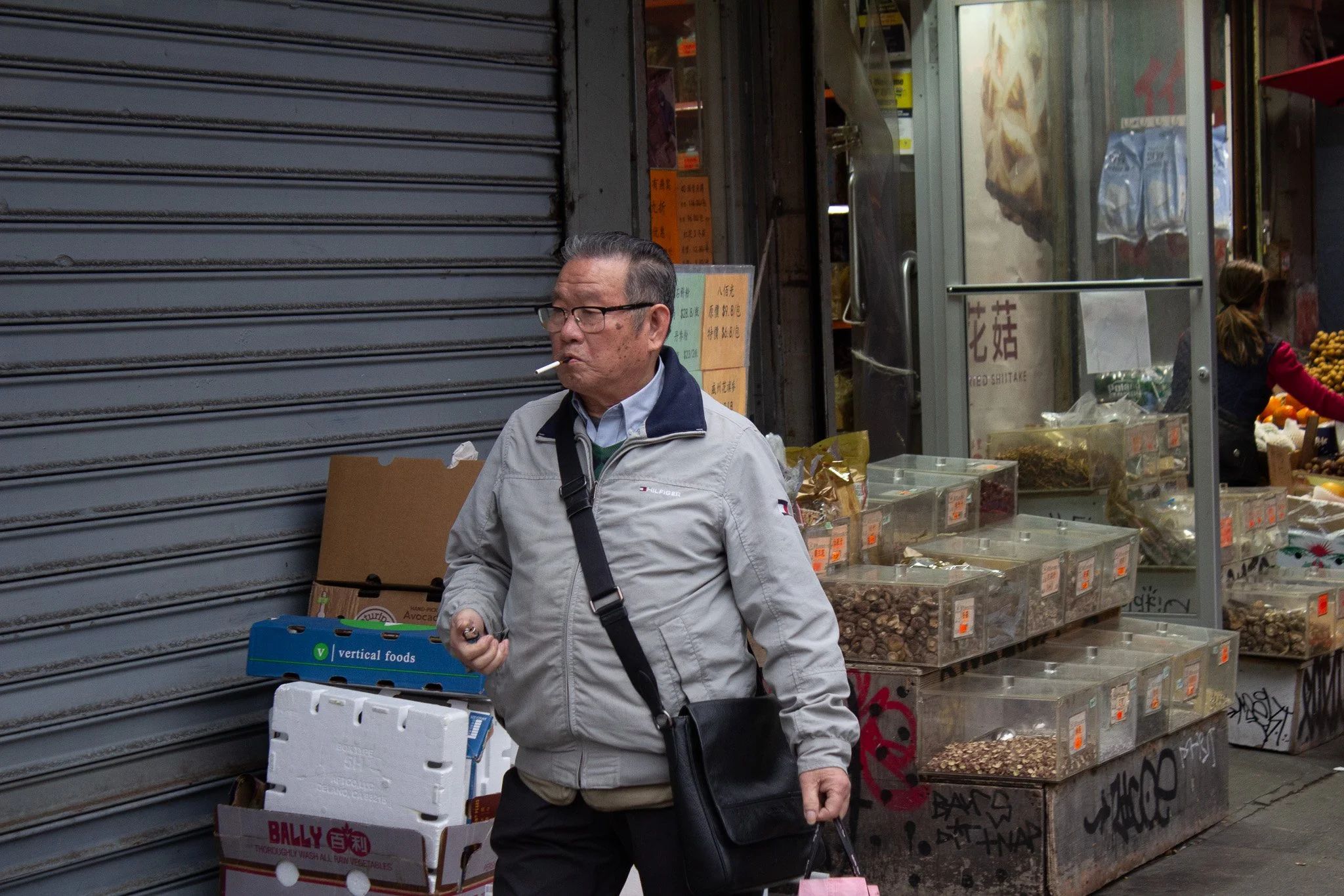 A man walking past a street market stall with dry goods in clear containers. The man is smoking a cigarette, wearing glasses, a light gray jacket, and carrying a black shoulder bag.