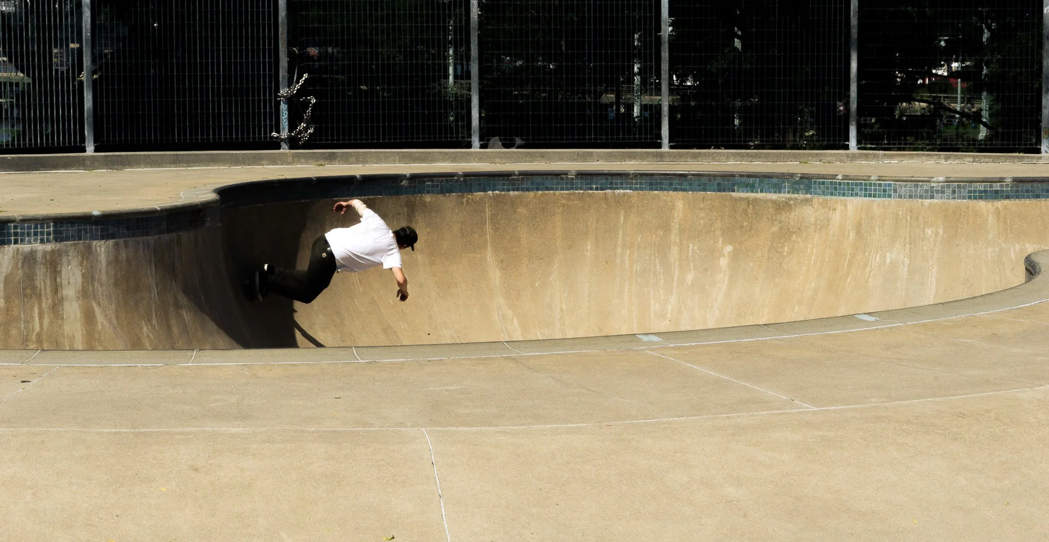 A man wearing a white shirt and black pants skating inside a concrete skatepark bowl, with a metal fence and trees in the background.