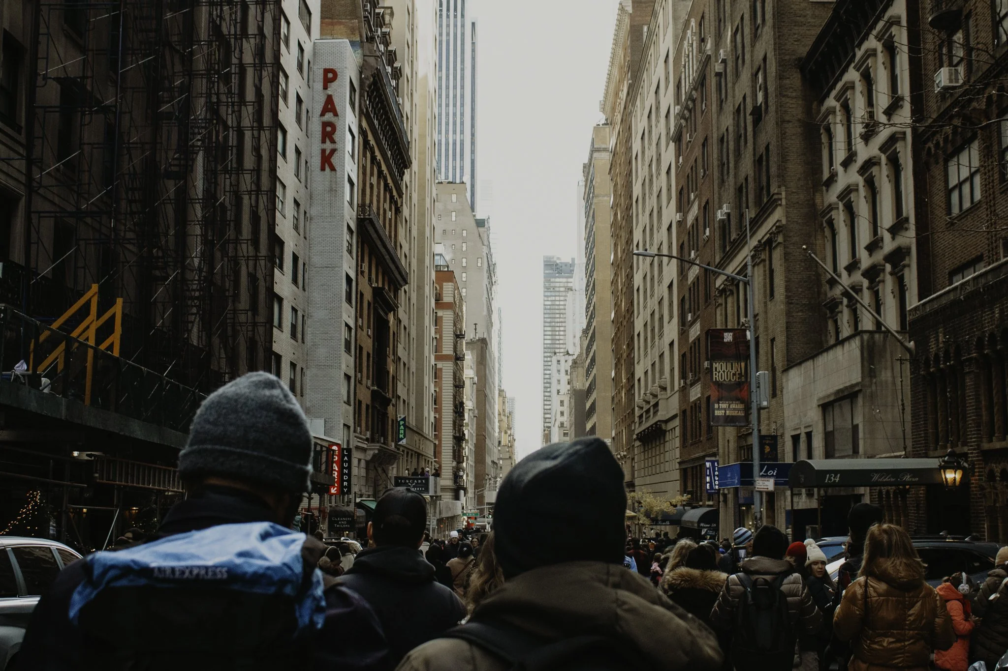 A busy city street full of pedestrians on a sidewalk lined with tall buildings, some with scaffolding, and a red parking sign on one building.
