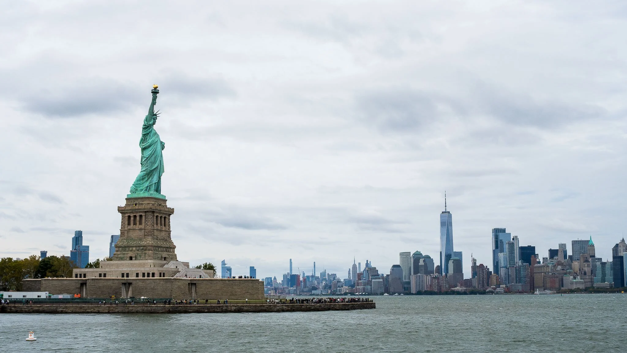 View of the Statue of Liberty on Liberty Island with New York City skyline in the background, featuring One World Trade Center and various skyscrapers, under a cloudy sky.