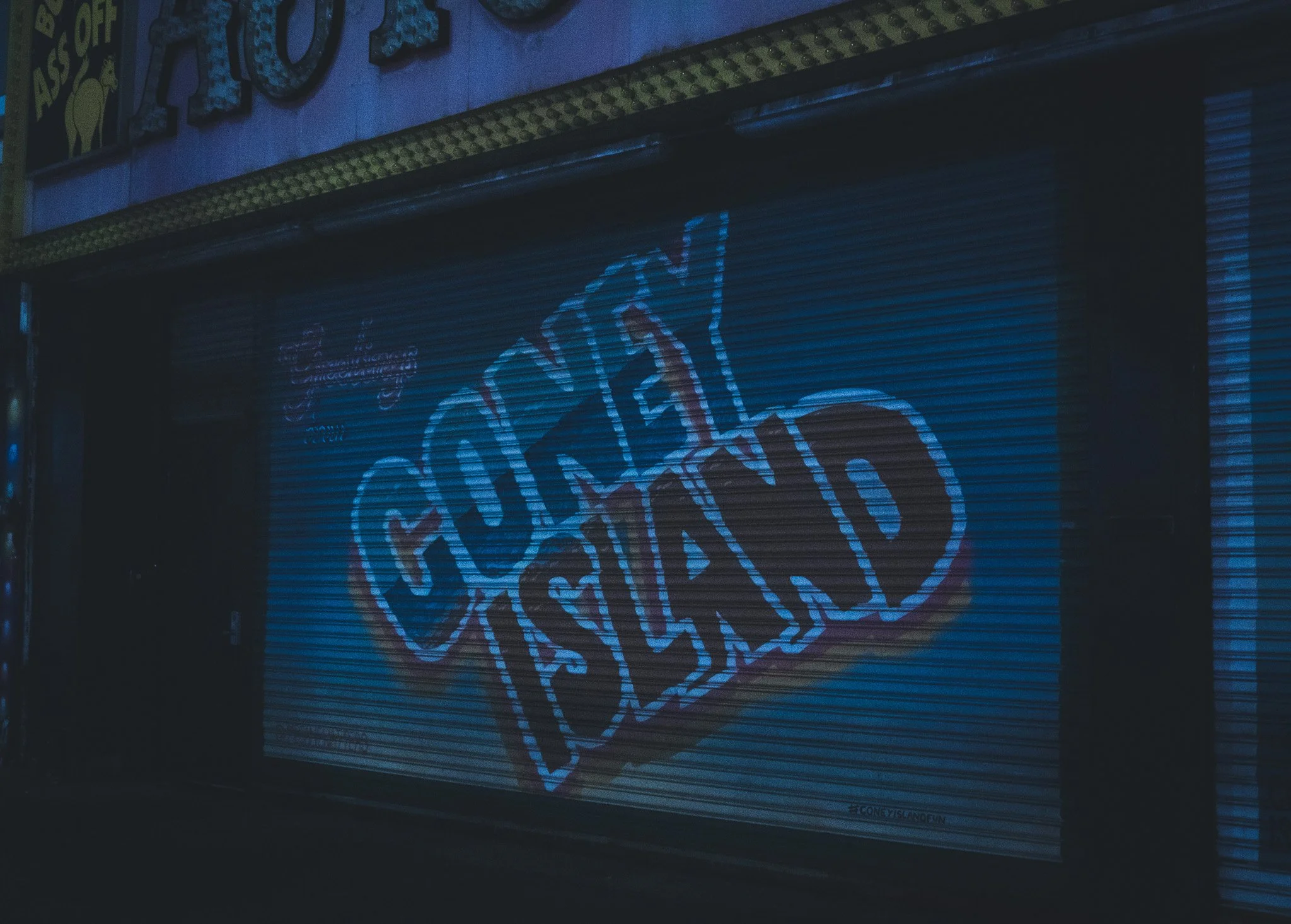 A dark street scene showing a closed metal shutter with a large, colorful graffiti heart design. Above the shutter, part of a building's wall is visible with some decorative molding.