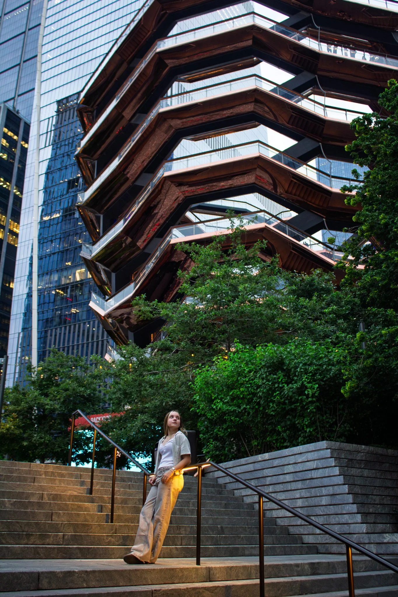 A woman standing on stairs at sunset near a tree in front of a modern building with an external escalator or walkway structure.