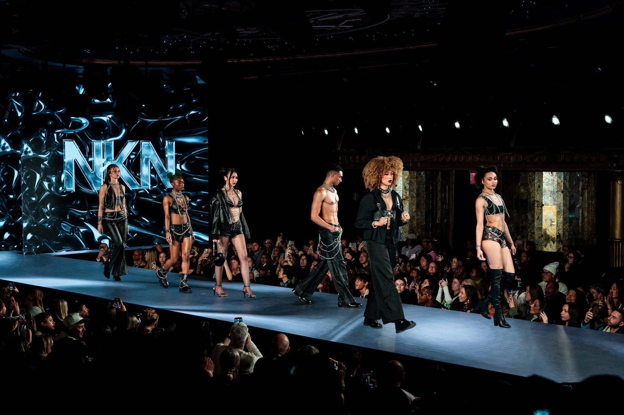 Models walk down a runway during a fashion show with an audience on both sides. The background features a decorative wall with the letters 'NN' illuminated.