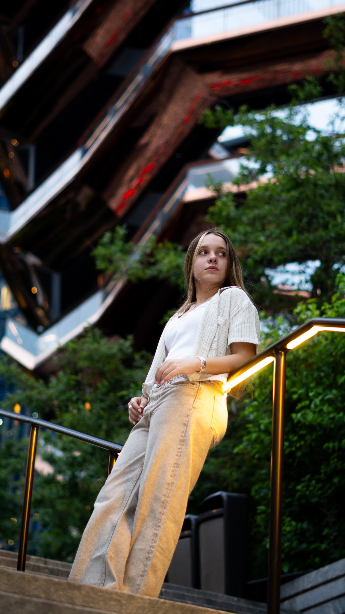 A young woman with long hair, wearing a light-colored blazer and beige pants, stands on a staircase with a black handrail, illuminated by a warm light, outdoors during the evening, with a background of green trees and an apartment building.