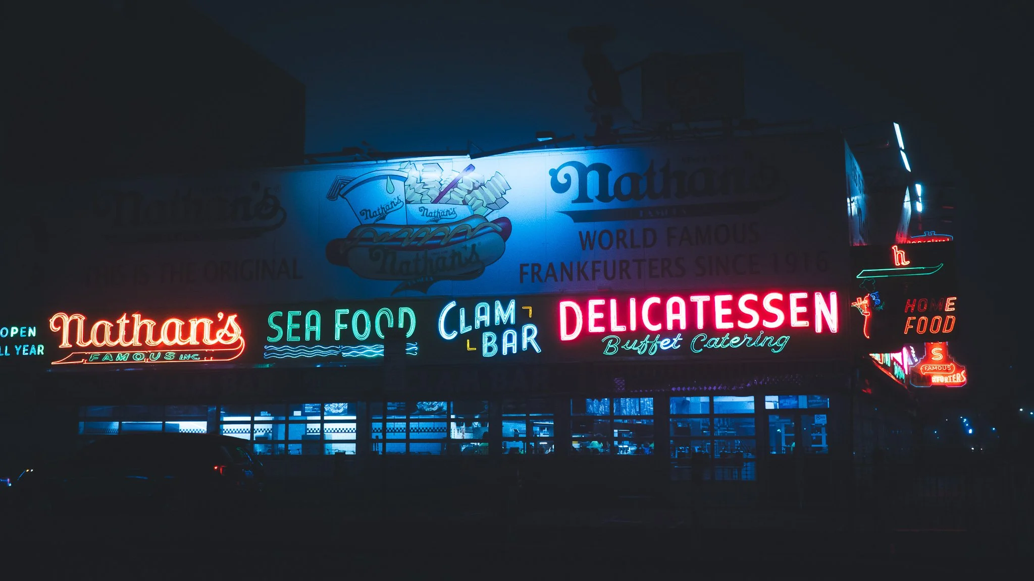 Night view of neon signs outside Nathan's Famous, a seafood and delicatessen restaurant. The signs advertise seafood, clam bar, buffet catering, and home food, with bright colors including red, green, blue, and white.