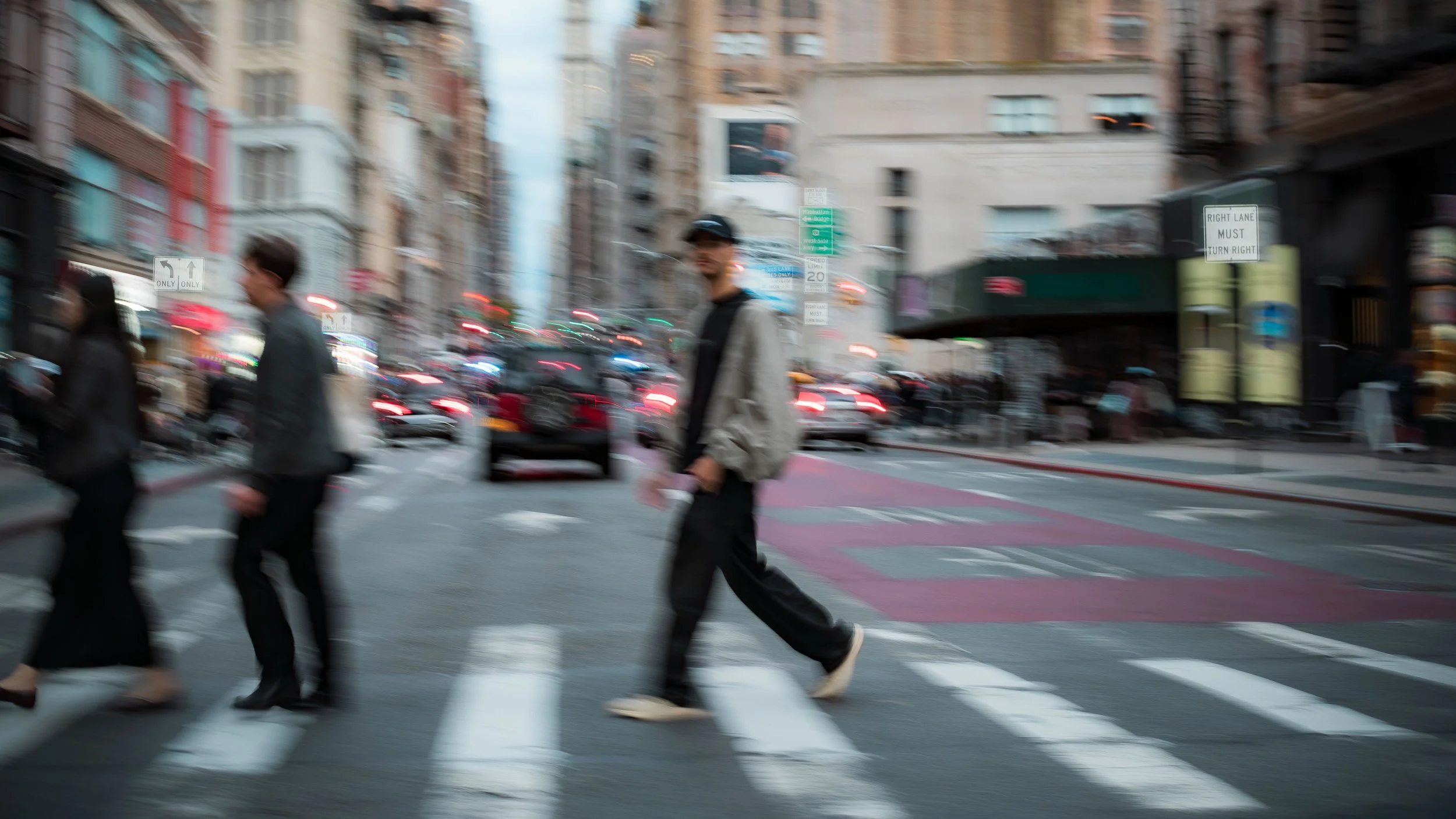 People crossing a busy city street during daytime, with cars and tall buildings in the background.
