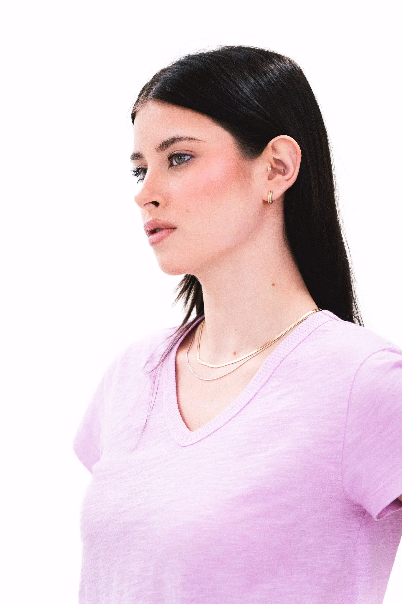 A woman with black hair, wearing gold hoop earrings, rings, and a pink V-neck top, looking to her left against a plain white background.