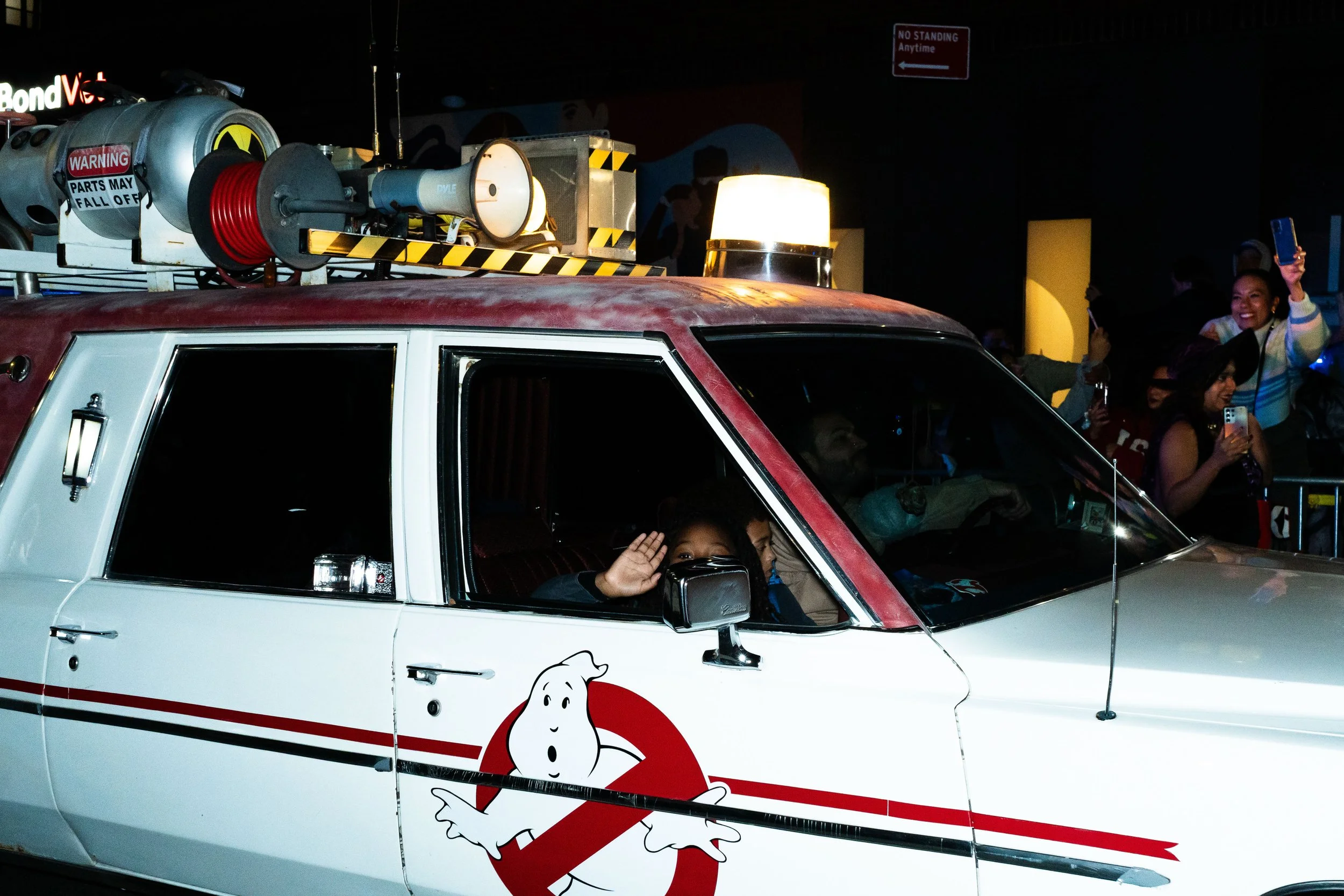 Children in a Ghostbusters-themed vehicle at a parade, with a crowd taking photos and waving nearby.