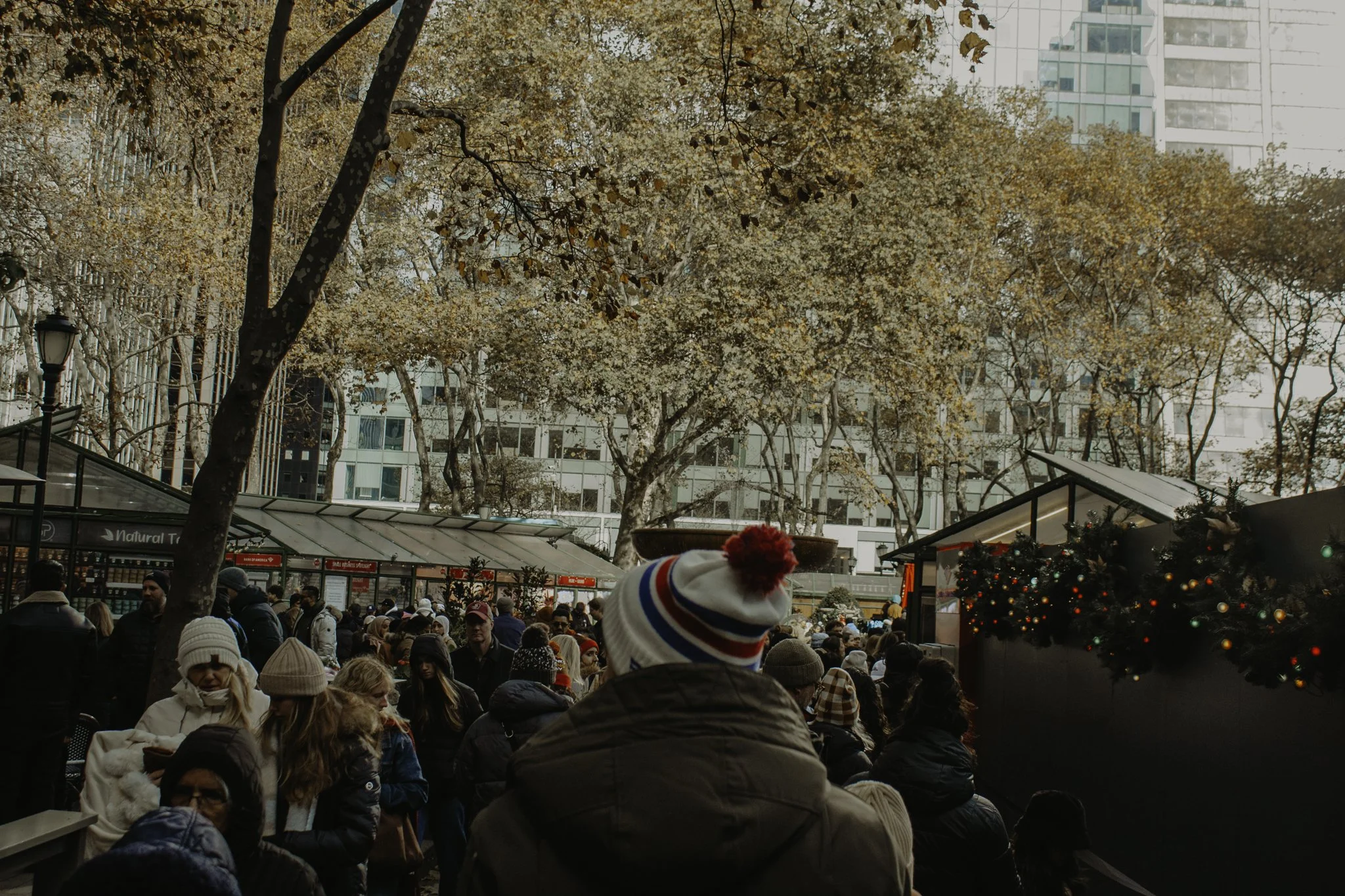 Crowd of people gathered outdoors during winter, with some wearing hats and coats. There are decorated Christmas trees and market stalls in the background, with trees that have autumn leaves, and tall buildings behind.