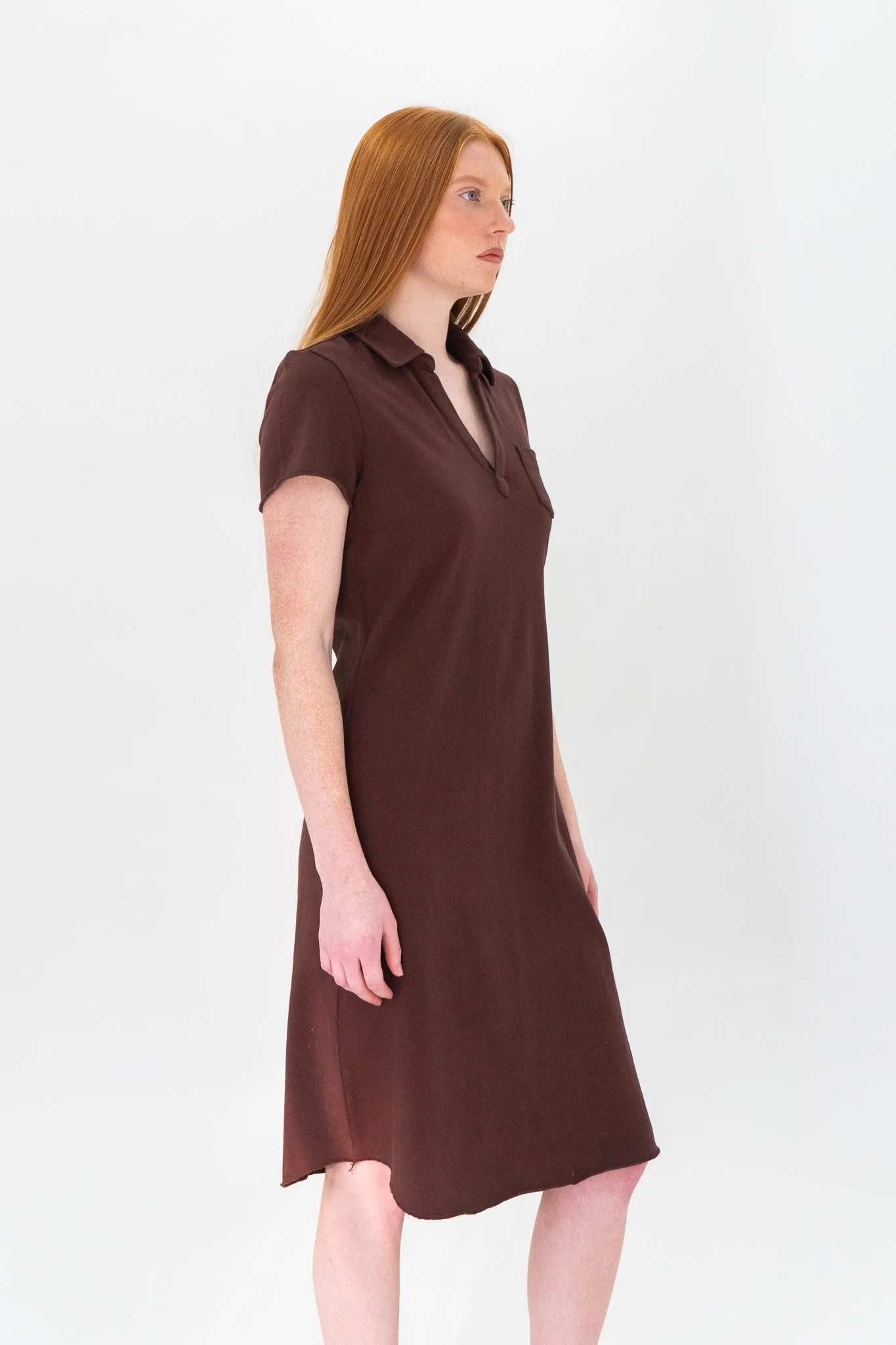 A woman with long red hair, wearing a dark brown dress, standing against a plain white background.