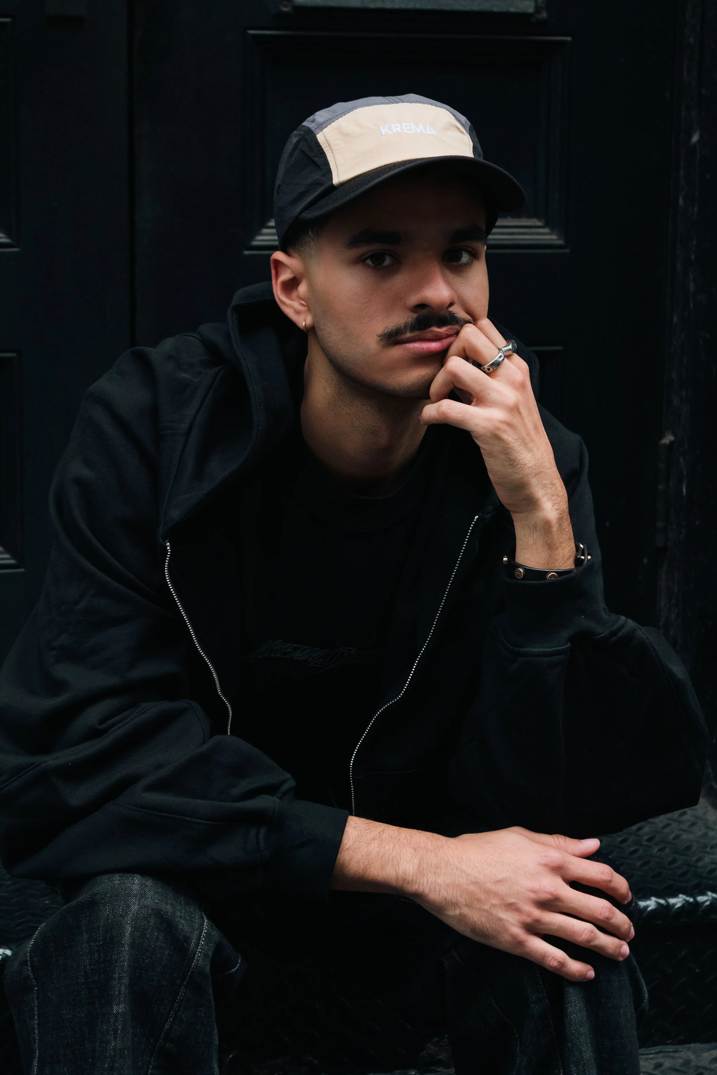 A young man with a mustache and dark eyebrows, wearing a black jacket and a beige and black cap, sitting against a dark background, resting his chin on his hand, looking at the camera.