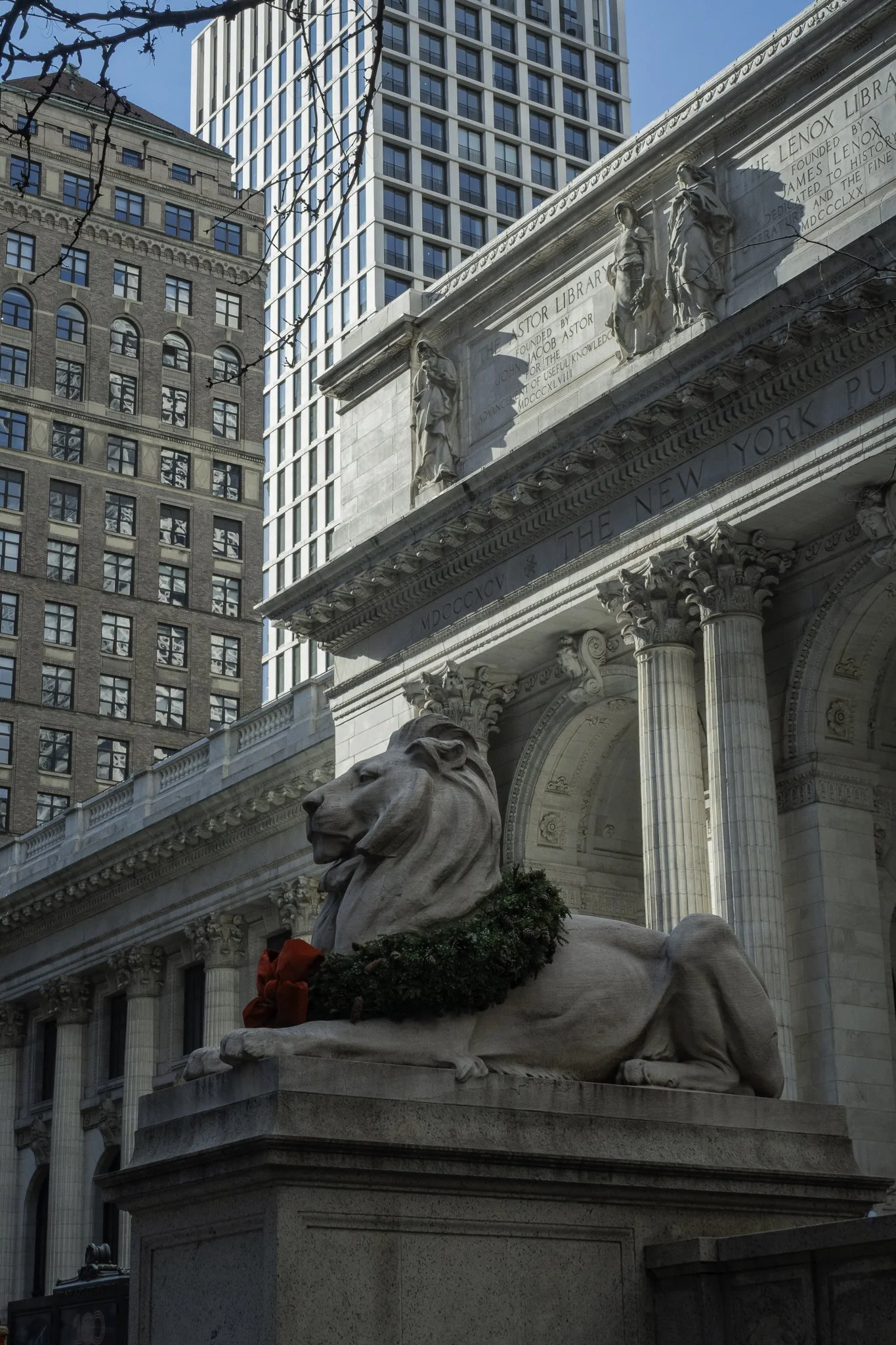 A stone lion statue with a wreath and red bow at the base, in front of a historic building with classical columns and detailed sculptures, surrounded by tall modern skyscrapers in New York City.