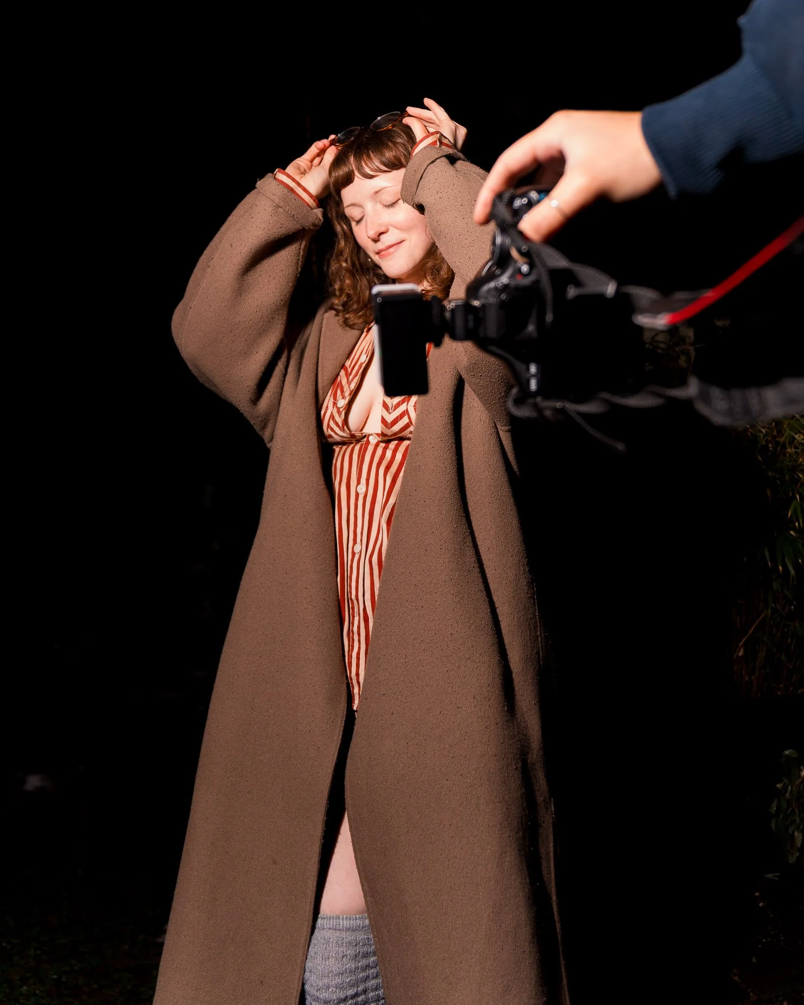A woman with curly hair and closed eyes smiling, wearing a beige coat over a striped shirt, with a person holding a camera nearby.