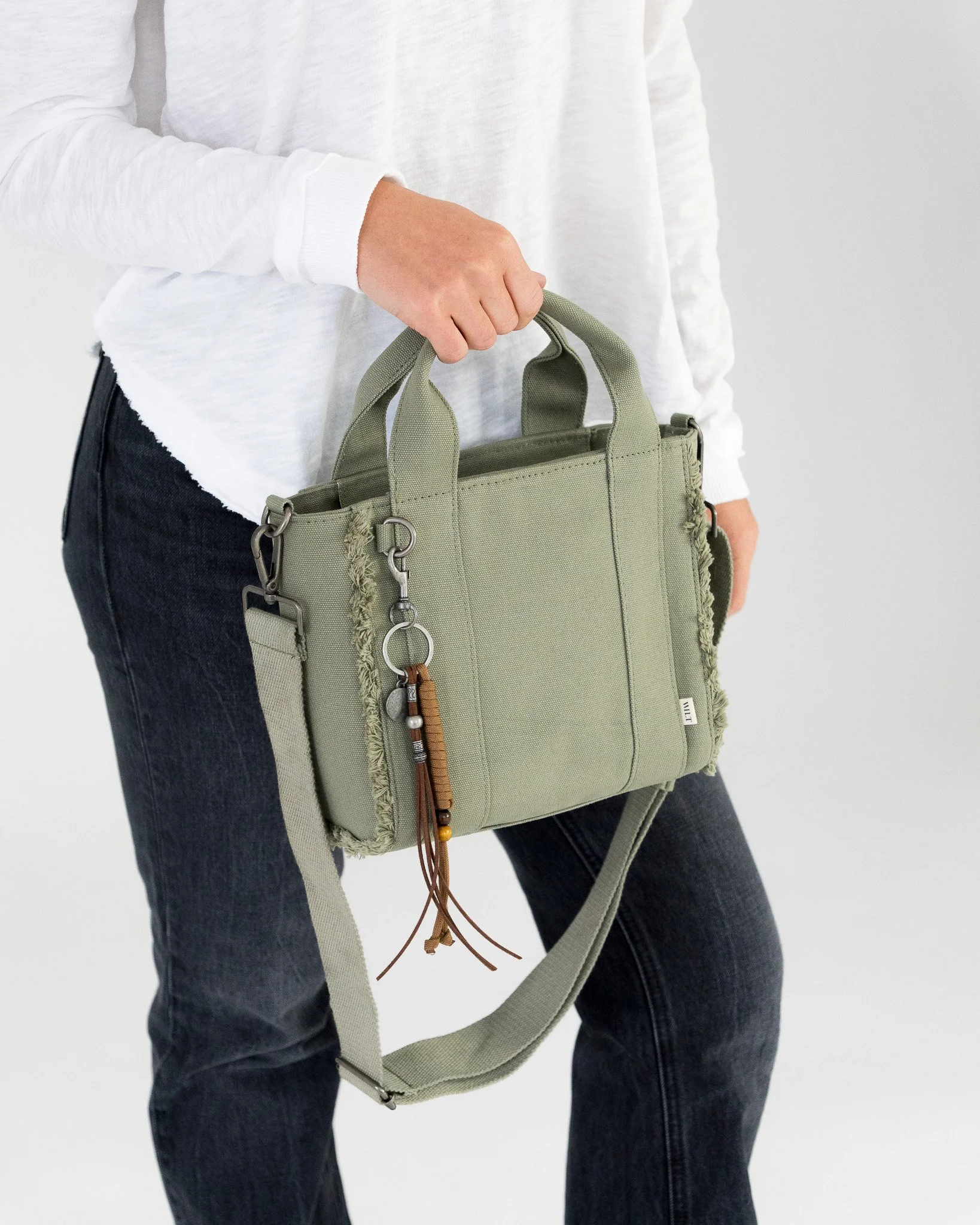 Close-up of a person wearing a white long sleeve shirt and dark jeans, holding an olive green handbag with key chains and decorative elements in front of a plain white background.