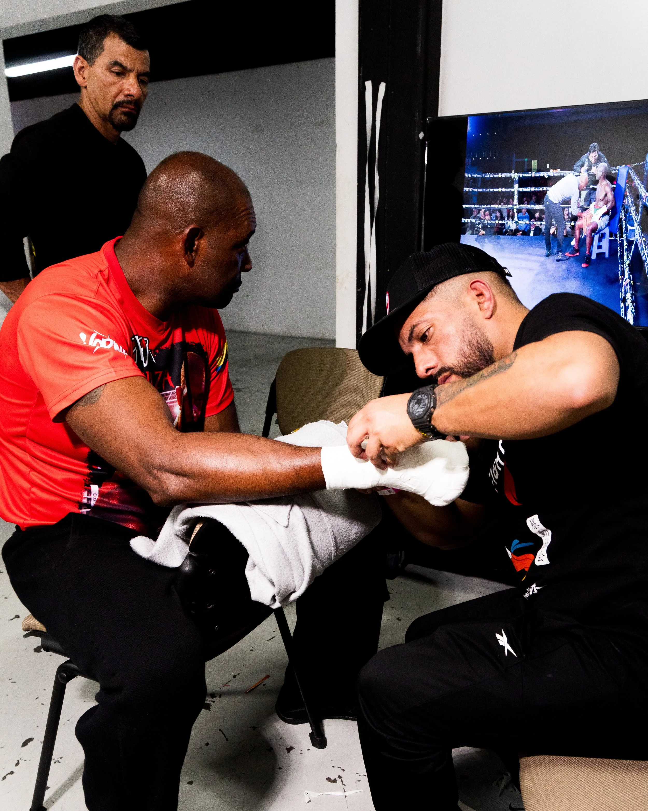 Two men are sitting at a table with one man appearing to receive medical treatment, while another stands beside him watching. There is a person in sports attire wearing a cap in the foreground, and a television screen in the background shows a boxing