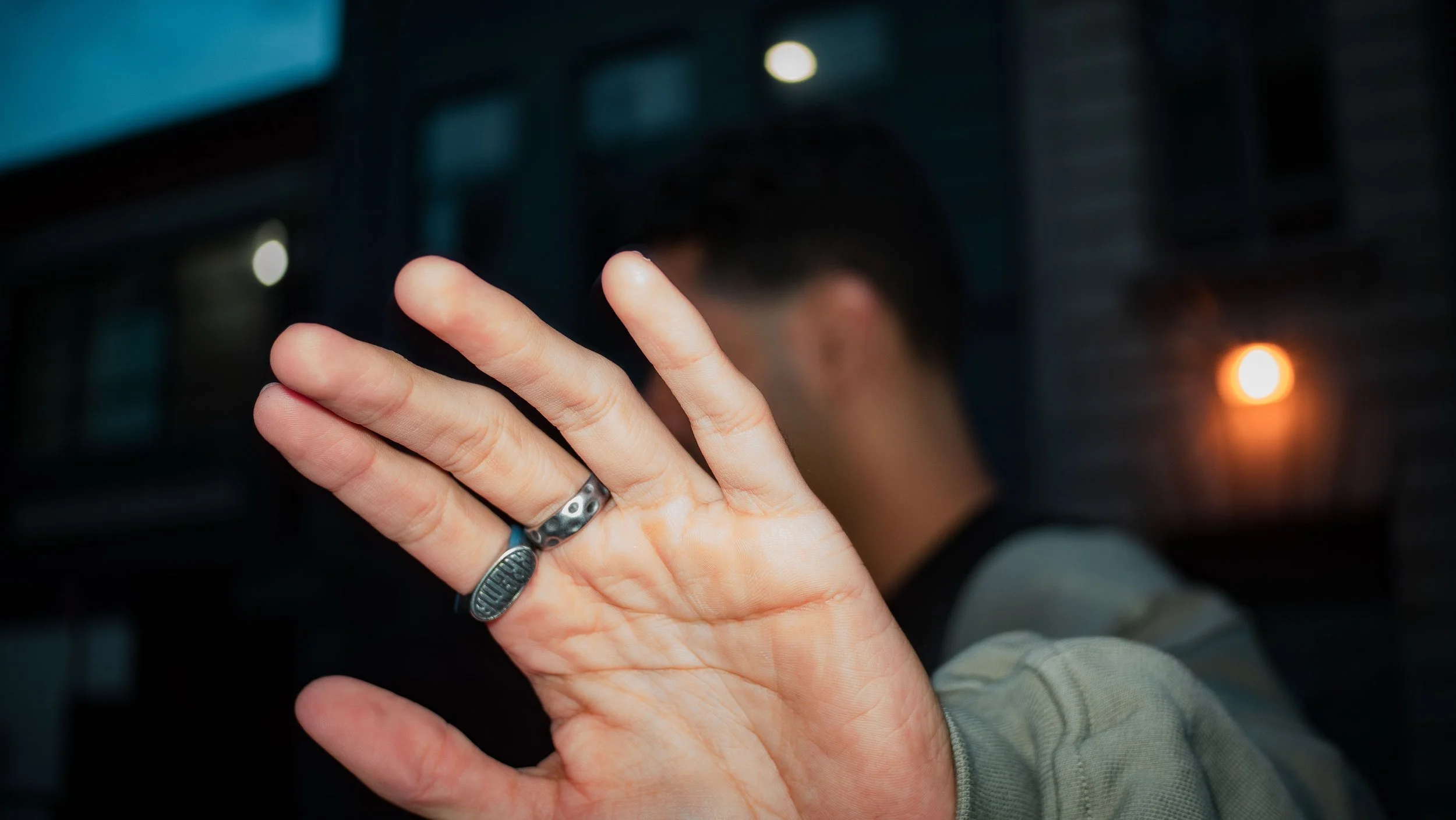 A person's hand raised with palm facing outward, wearing two rings, with a blurred background of a man's profile and city lights at night.
