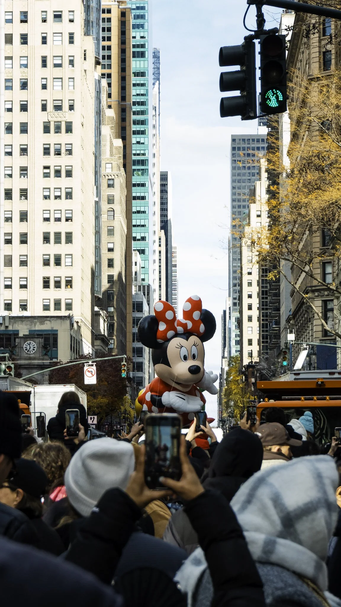A parade on a city street featuring a float with a large Minnie Mouse figure, surrounded by spectators taking photos, with tall buildings in the background and a green pedestrian traffic light.