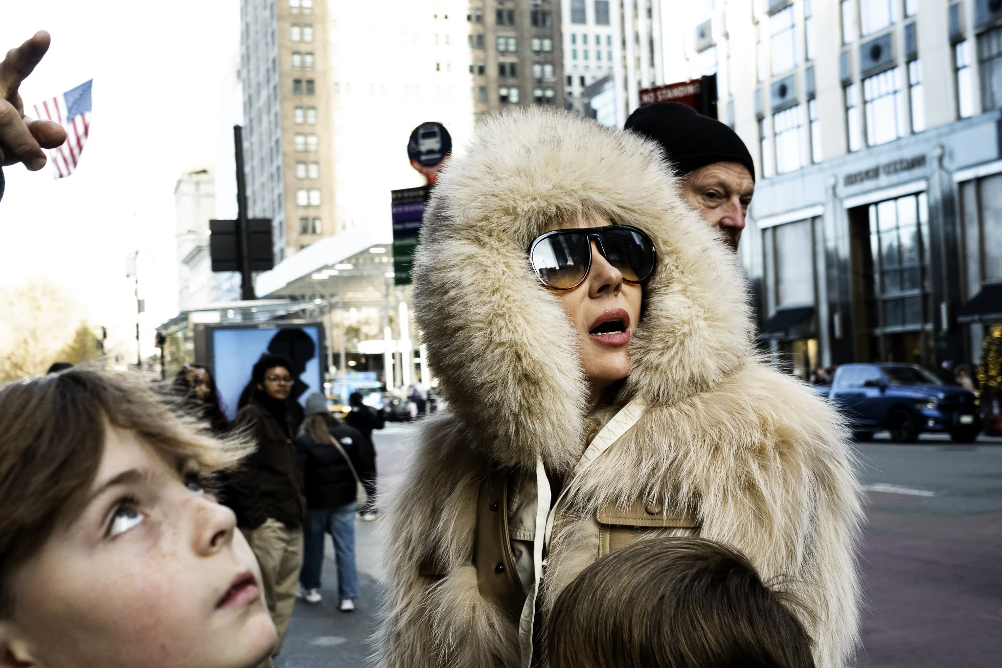 A woman wearing sunglasses and a large furry hooded coat standing among children on a city street, with tall buildings and other pedestrians in the background.
