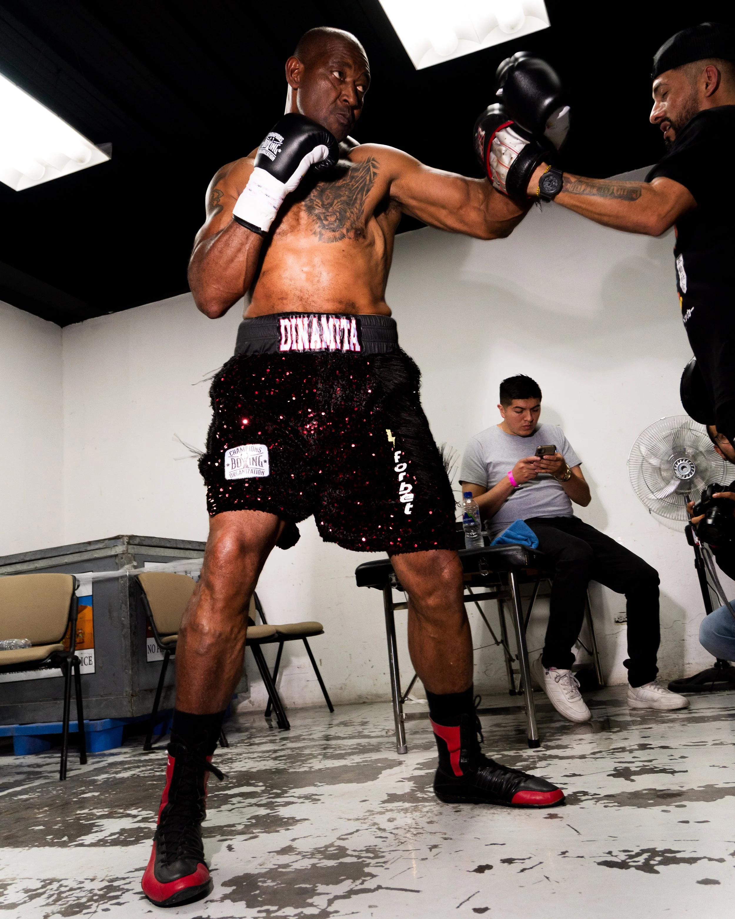 A shirtless man with tattoos sparring with a trainer in a boxing gym. The man is wearing black and red boxing shorts and black boxing gloves, and is throwing a punch. The trainer is holding pads to catch the punch. Two seated individuals are in the b
