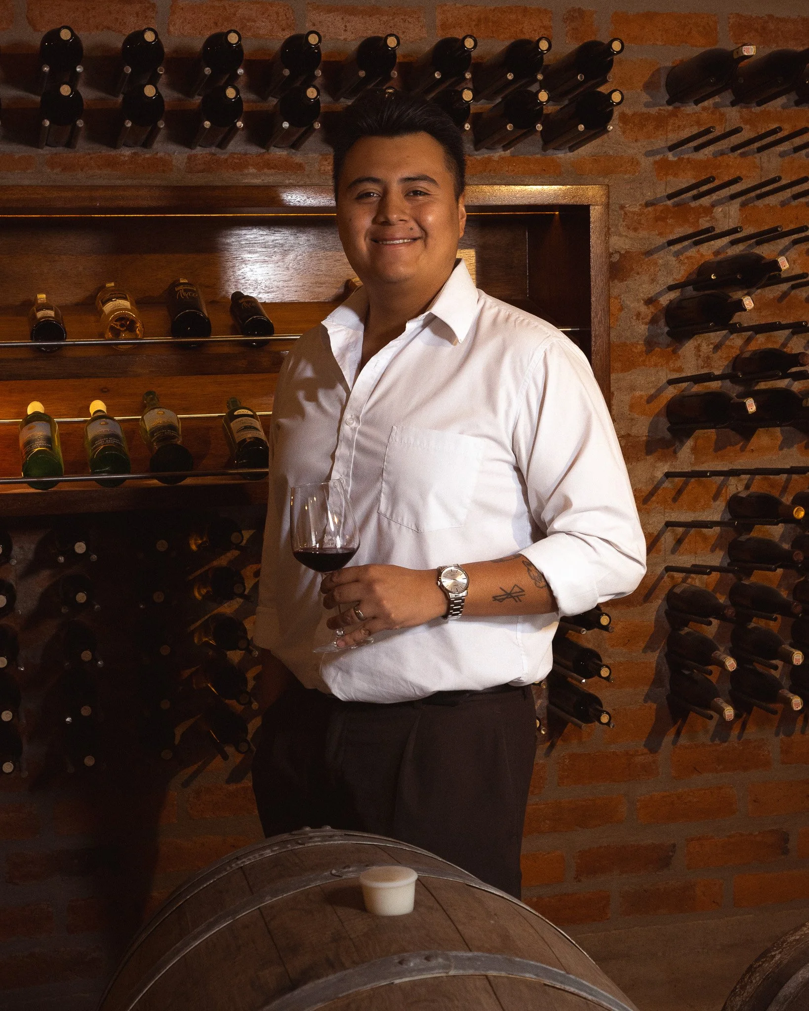 A man with dark hair styled back, wearing a white button-up shirt and a wristwatch, standing in a wine cellar with wine bottles stored on brick walls and wooden racks, holding a glass of red wine.