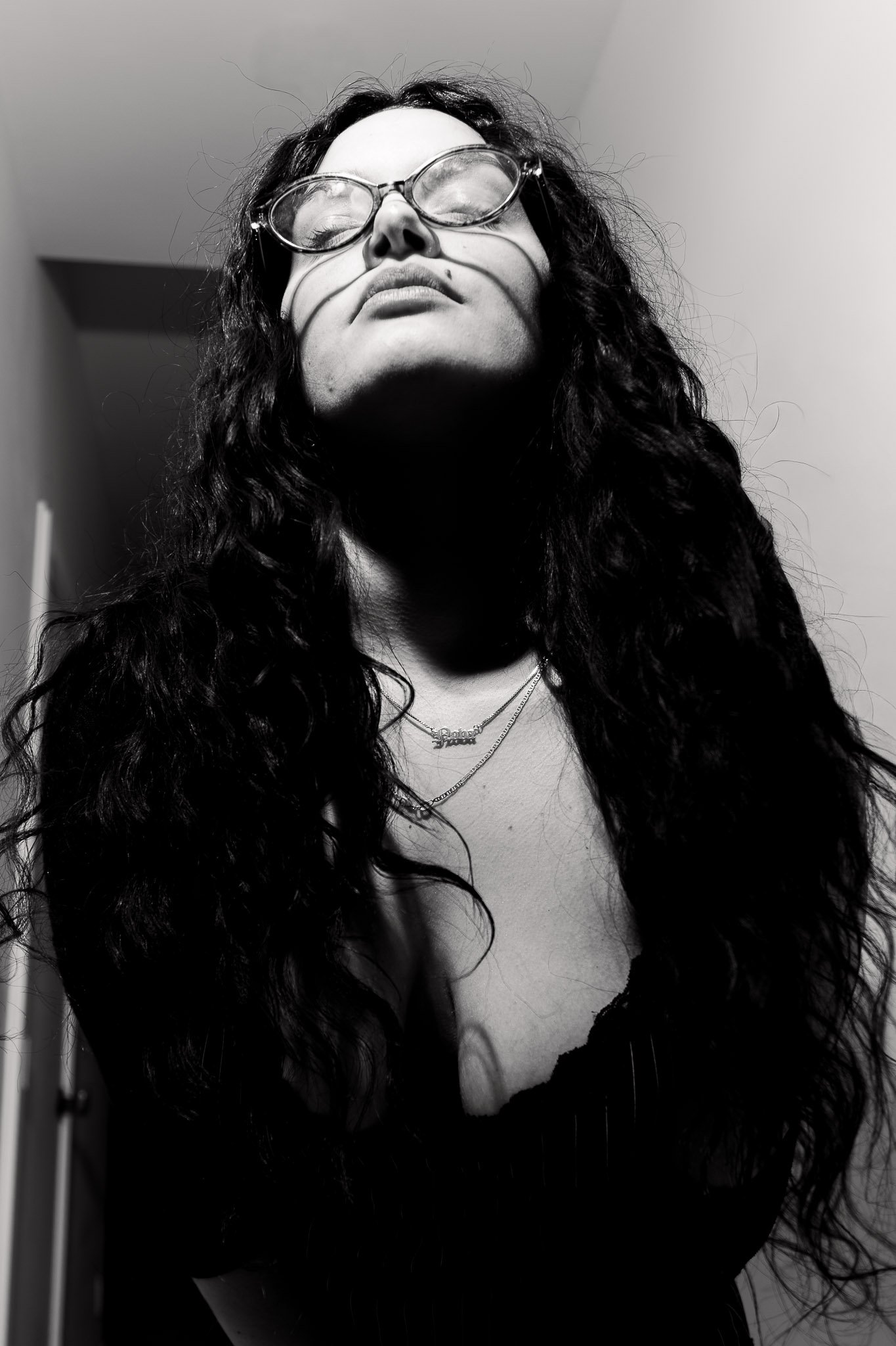 Black and white photo of a woman with long, curly hair, wearing glasses and a necklace, posing with her head tilted back and eyes closed.