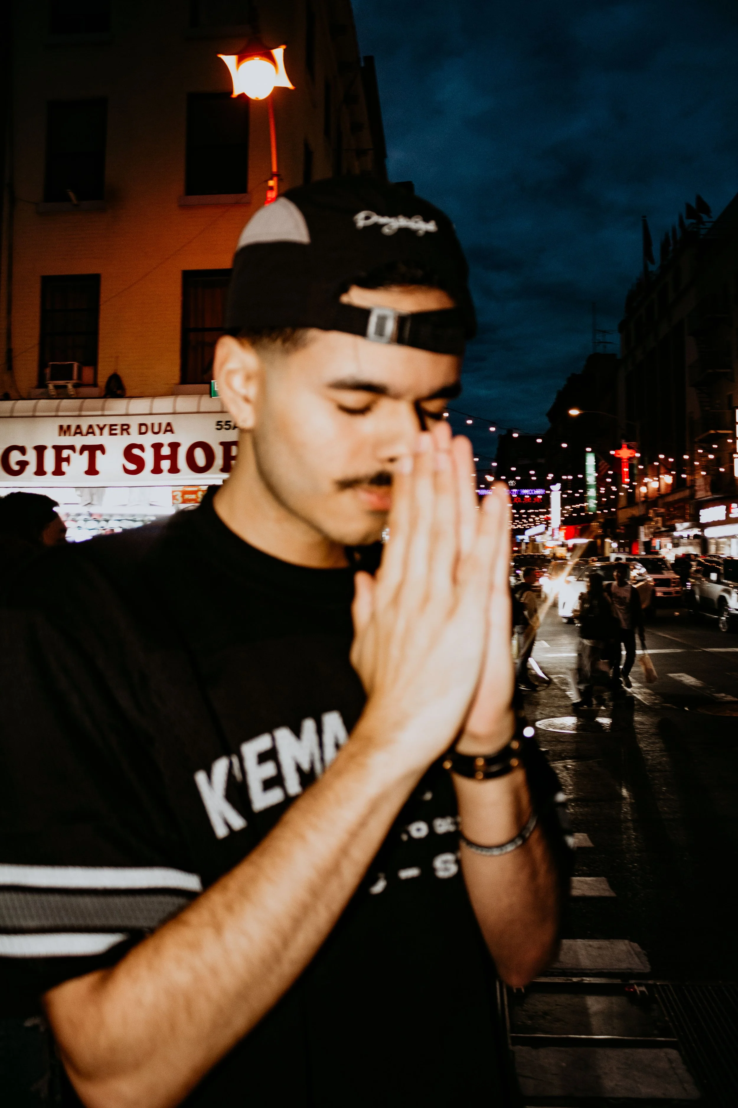 A young man with a beard and mustache wearing a black cap backwards and a black t-shirt, standing on a busy city street at night with his eyes closed and hands pressed together in prayer or meditation.