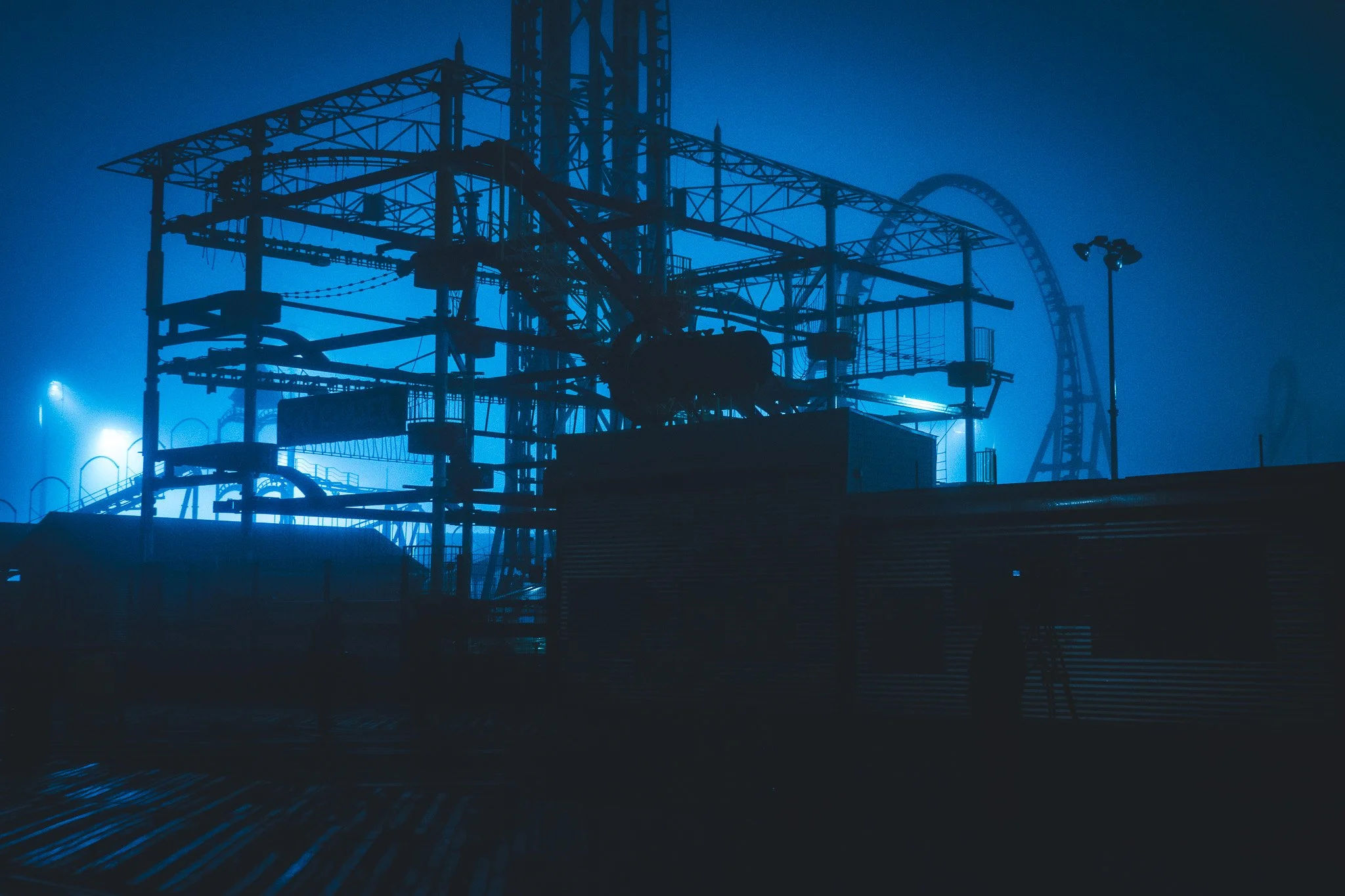 A silhouetted amusement park roller coaster at night with blue lighting, visible roller coaster tracks and supporting structures.