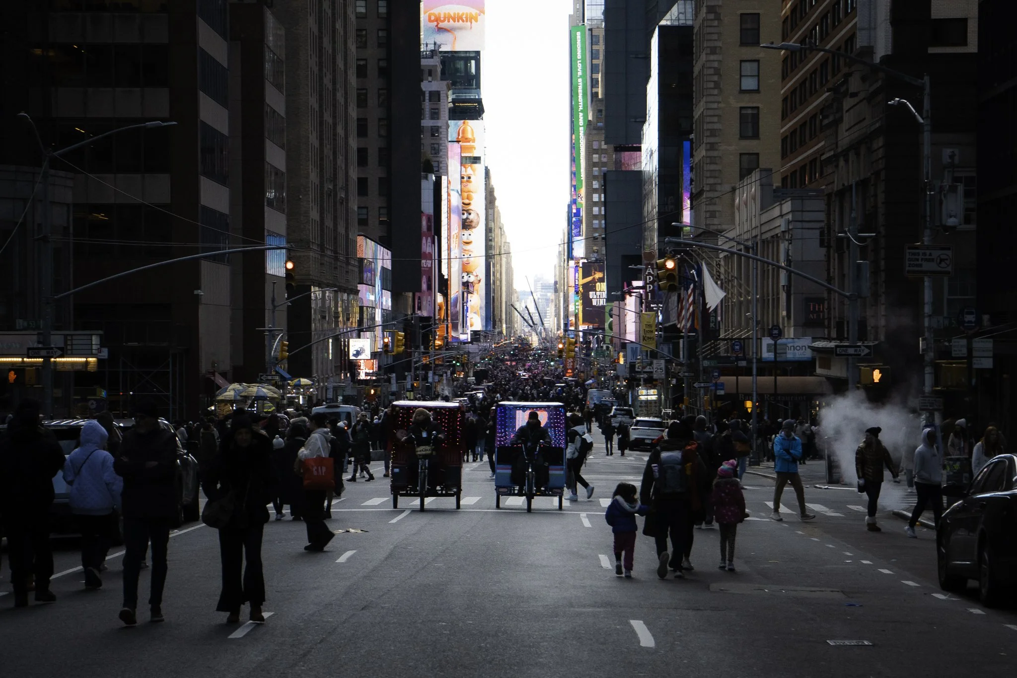 Busy city street filled with pedestrians, some on bicycles, with tall buildings on both sides and electronic billboards overhead.