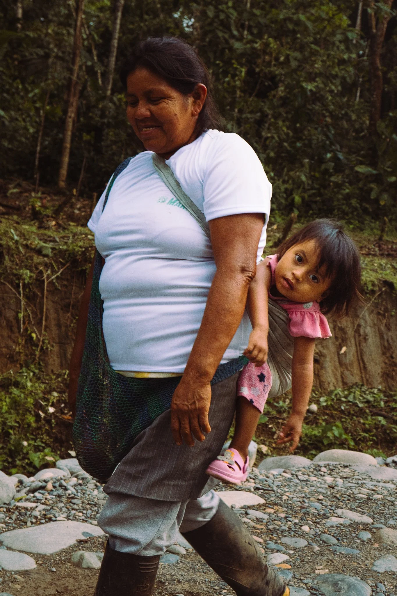 An elderly woman and a young girl are outdoors in a forested area. The woman is smiling and wearing a white shirt, with a backpack strap over her shoulder. The girl, who is leaning on the woman's side, has long dark hair, pink shoes, and a pink outfi