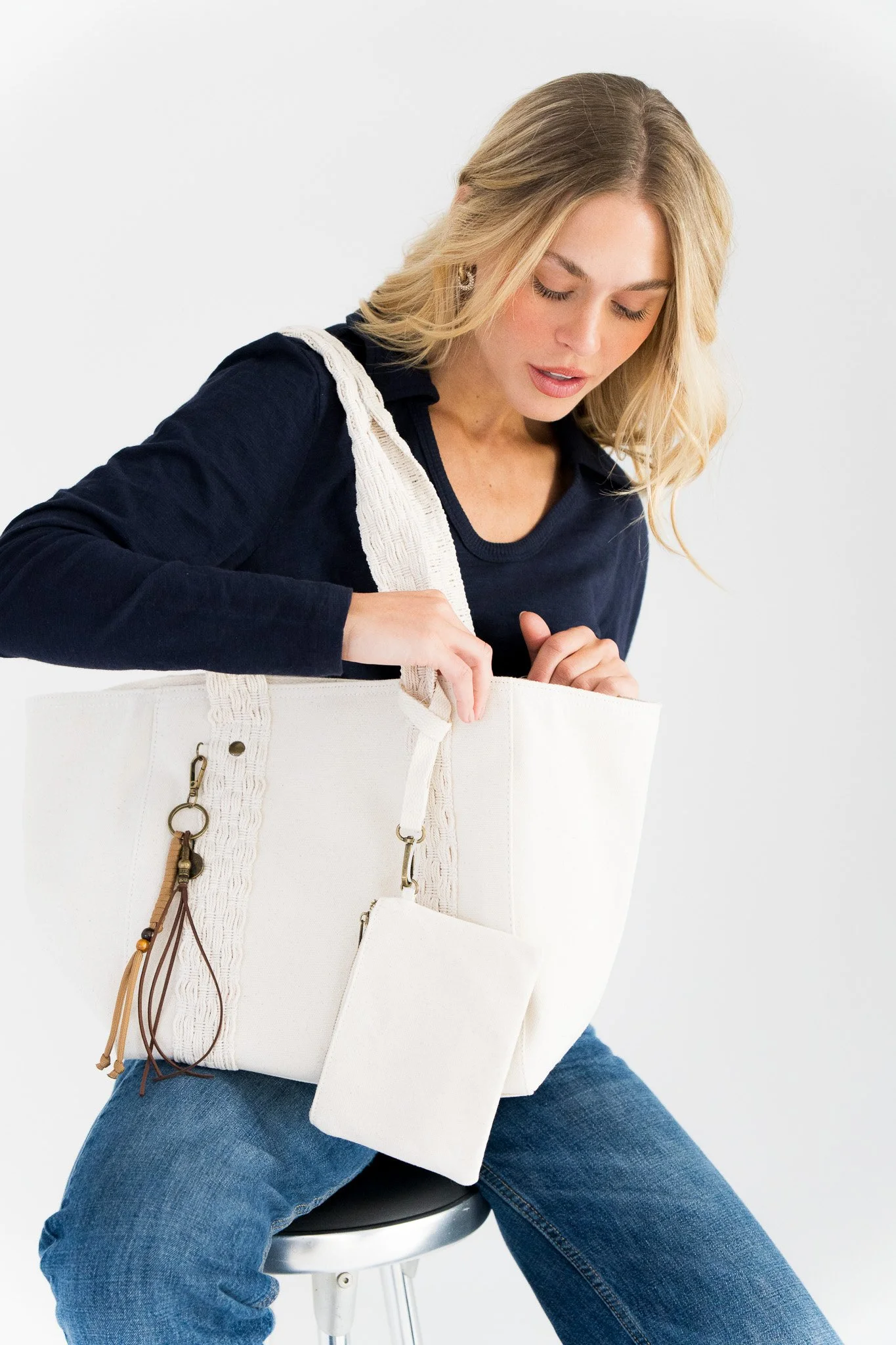 A woman in a navy shirt and jeans sitting on a stool, looking into a large white tote bag with decorative straps, against a plain white background.