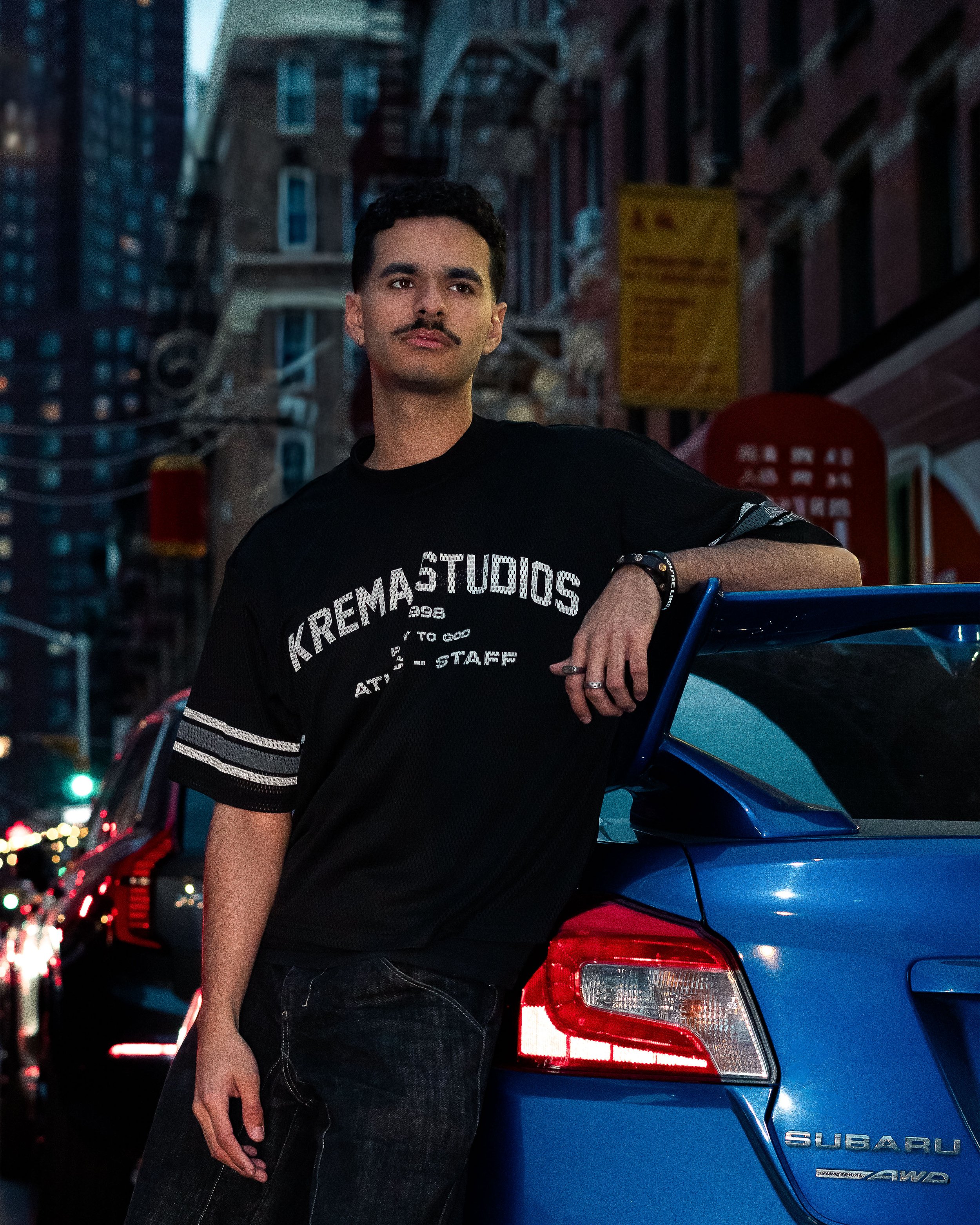 A young man with a mustache and short dark hair leans against a blue Subaru car in an urban street scene during dusk, wearing a black T-shirt with white lettering and dark jeans.