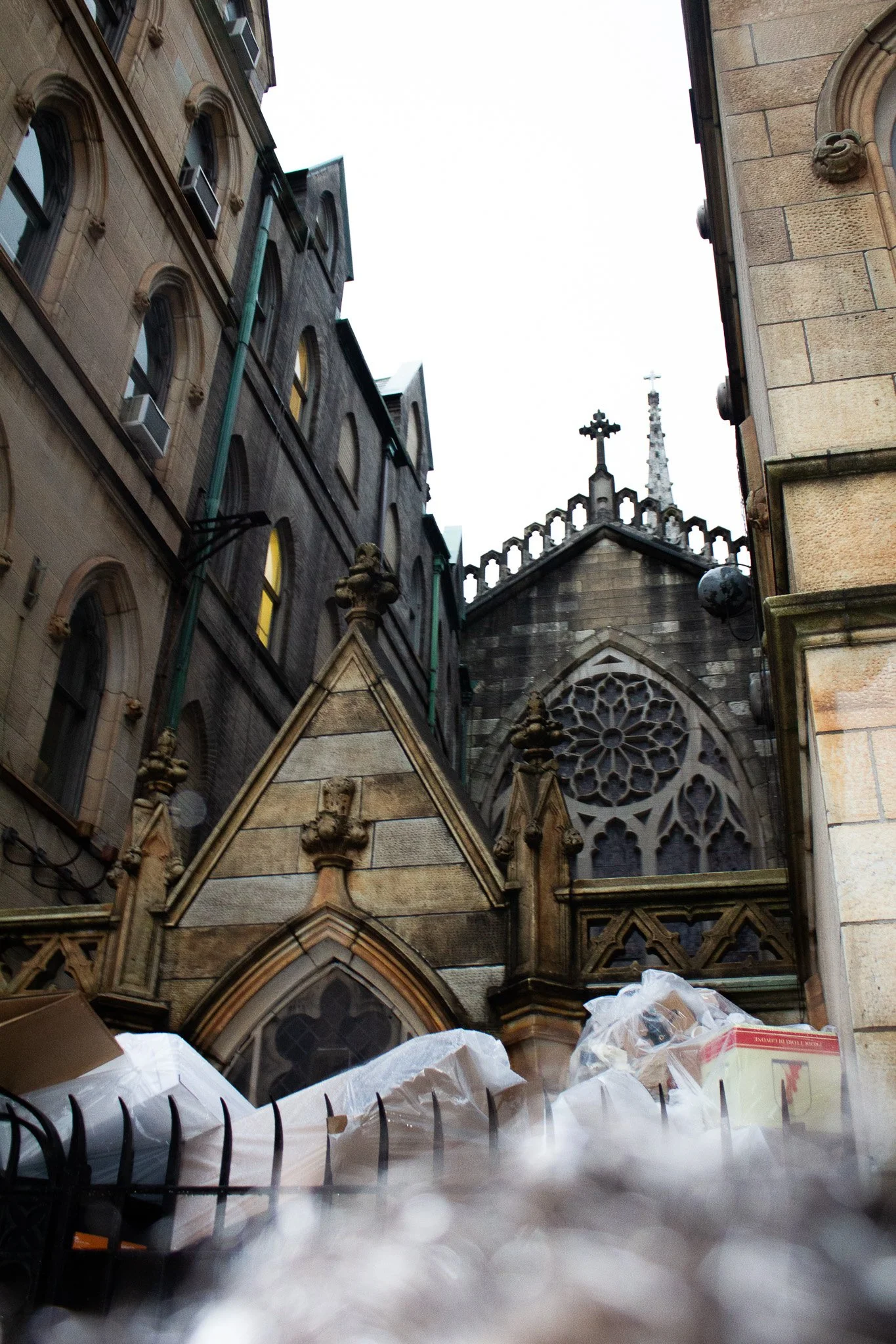 View of a Gothic church with stained glass rose window, surrounded by tall stone buildings, seen from a low angle, with some construction materials and a fence in the foreground.