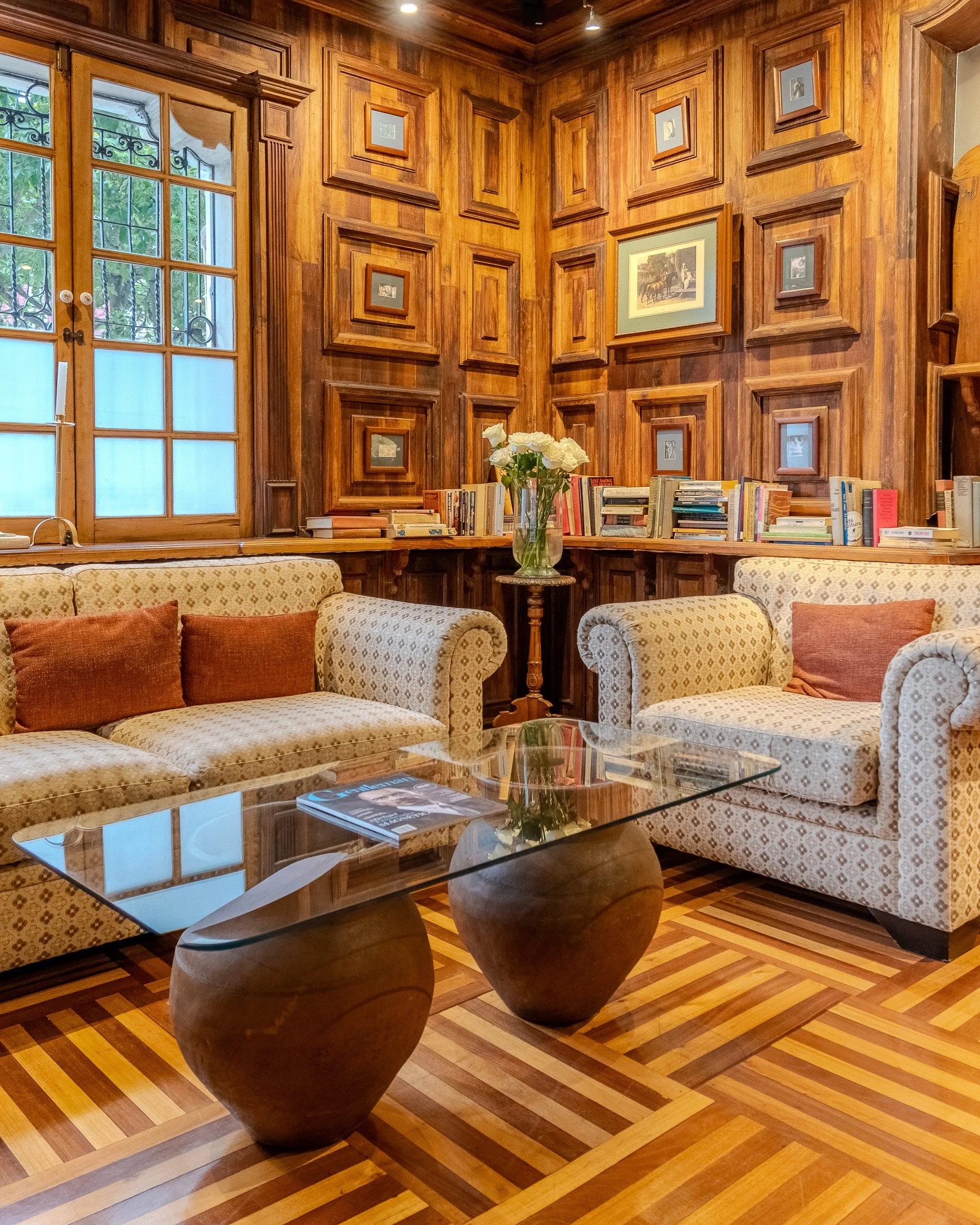 A cozy interior room with wood-paneled walls, framed artwork, a glass coffee table, and upholstered sofas with throw pillows. A large window with decorative ironwork reveals greenery outside. A vase of white flowers sits on a side table among books.