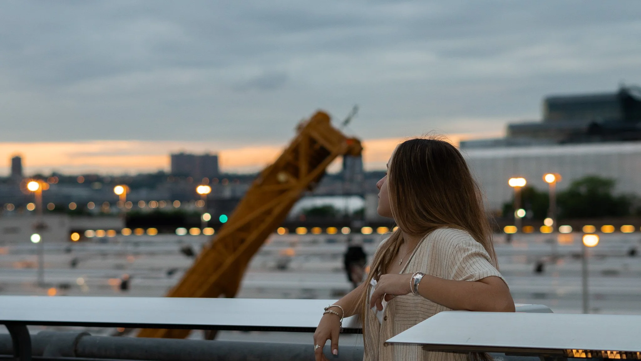 A woman with long brown hair sitting outdoors at dusk, looking to her right, with city buildings and lights in the background.