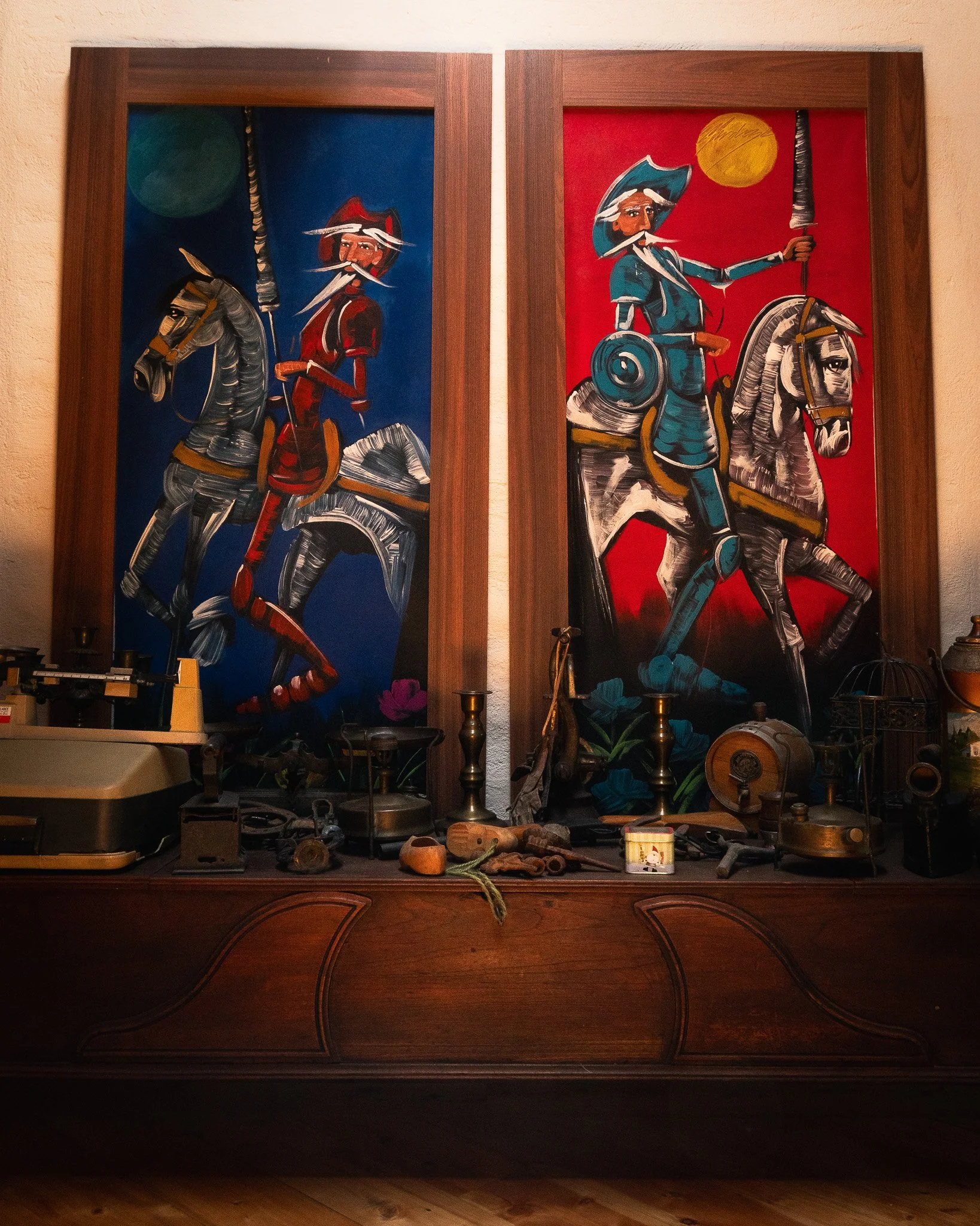 A wooden desk with vintage objects, including a typewriter, a drum, a lantern, and pottery, with two large colorful paintings of cowboys riding horses hanging above the desk.