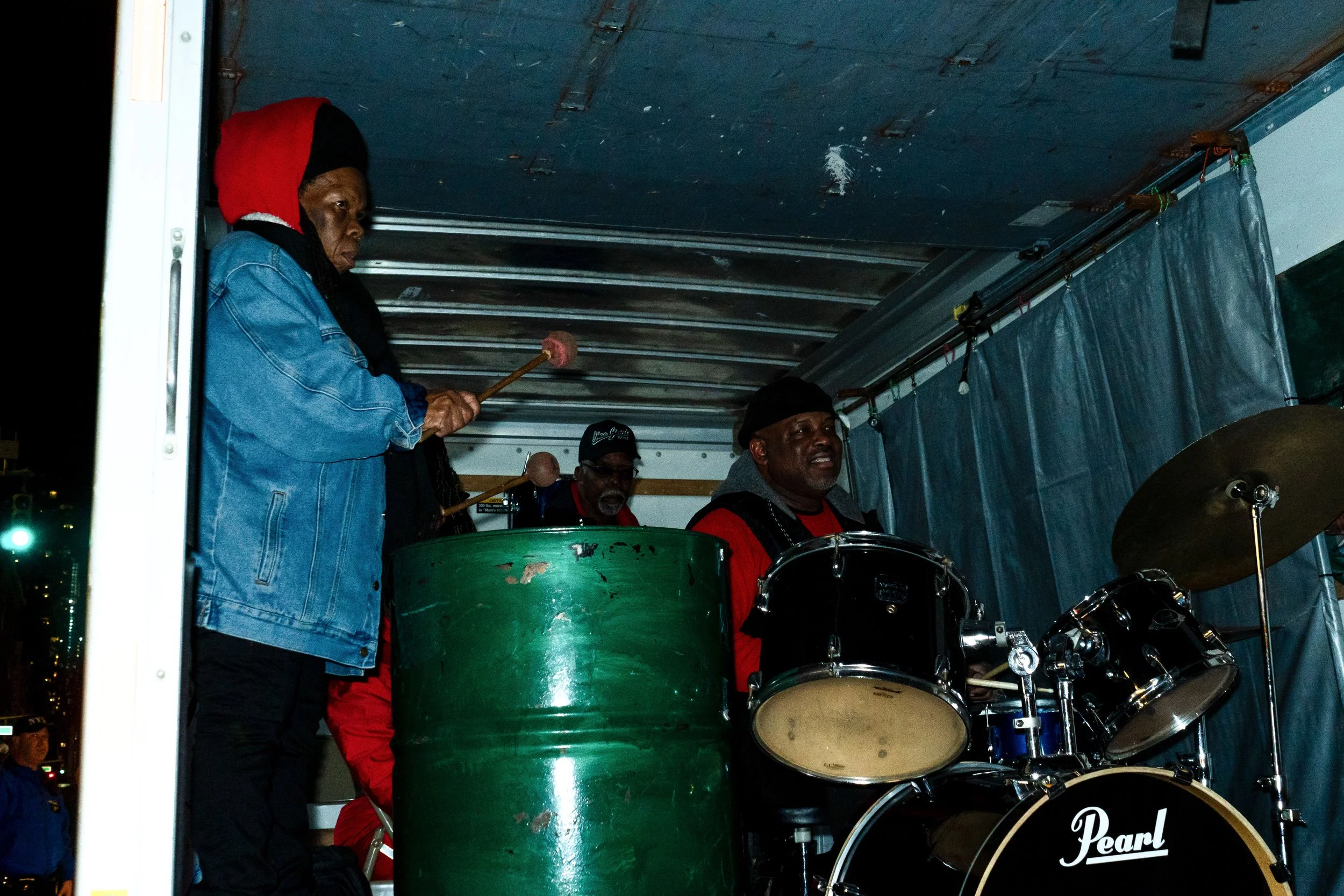 Three musicians performing in a truck, one woman playing a percussion instrument and two men playing drums.