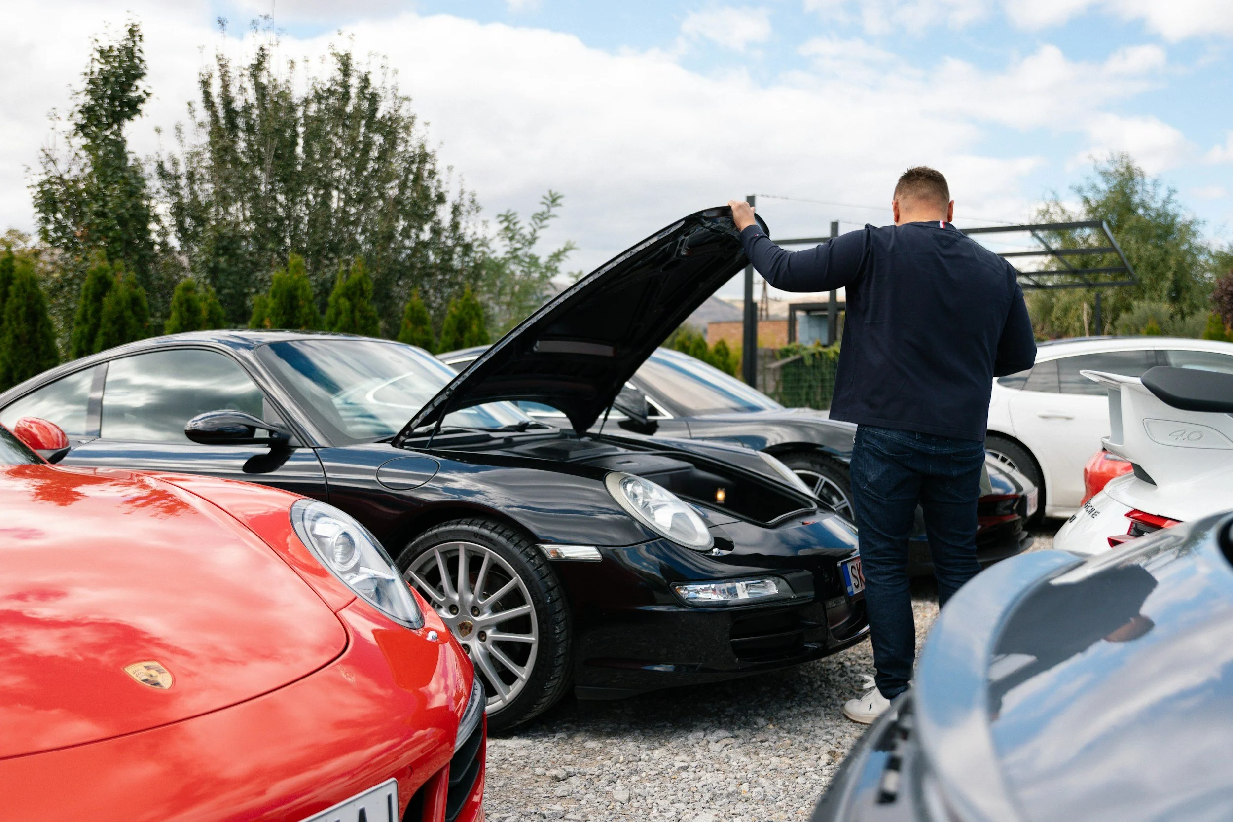 A man in a dark jacket inspecting a black Porsche with its hood open, parked in a lot surrounded by other cars and greenery.