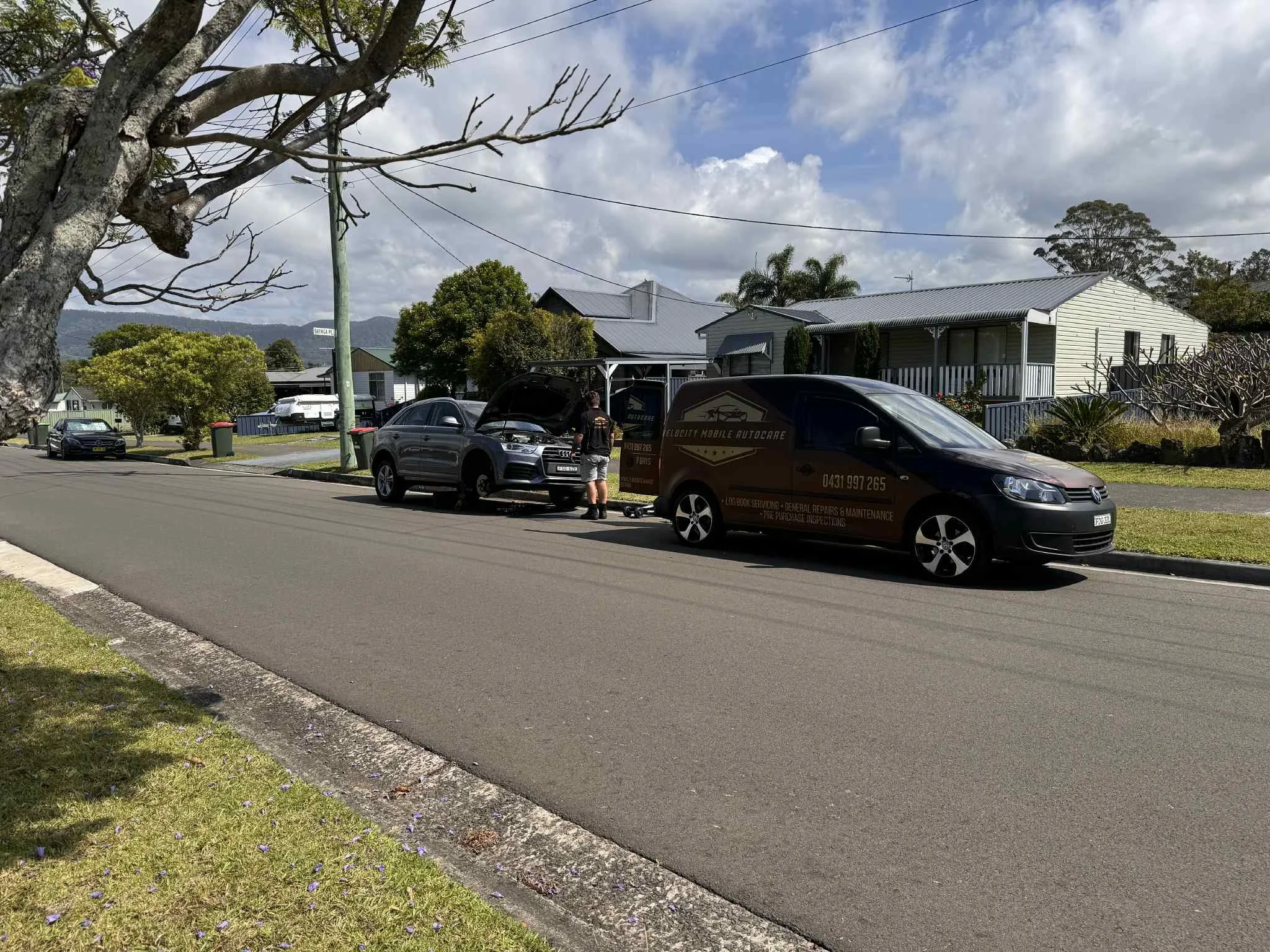 A man working on a car with its hood open parked on a residential street, near a branded auto repair van.