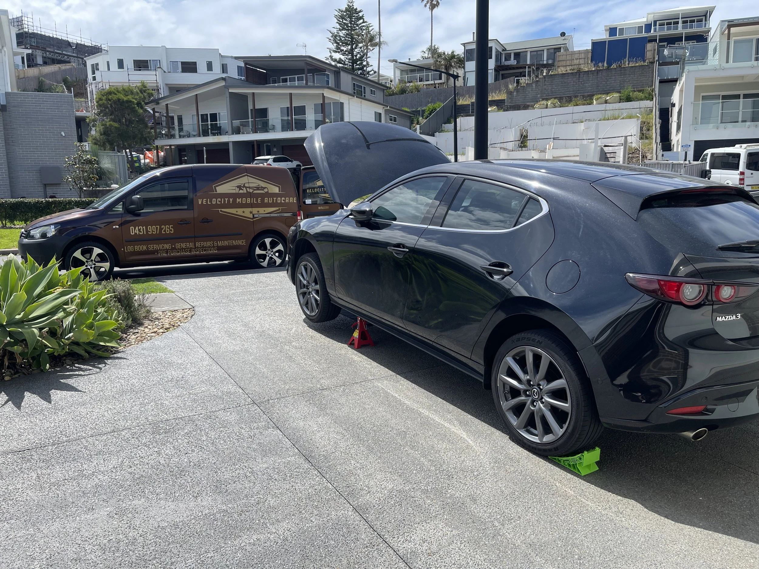 A black Mazda 3 car with a raised hood and green wheel chocks, parked on a driveway next to a brown Velocity Mobile Auto Care van with an open hood, in a residential area with modern houses in the background.