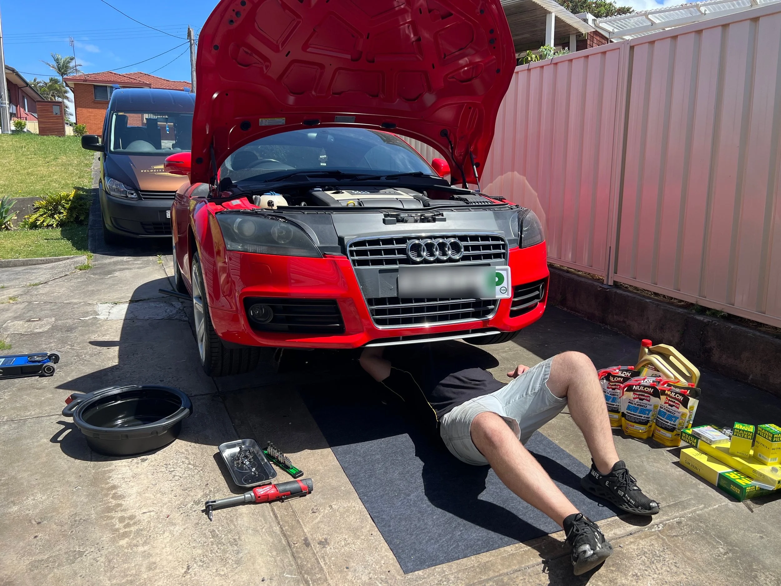 A person lying on the ground working under the hood of a red Audi car parked in a driveway, with car maintenance supplies and tools around.