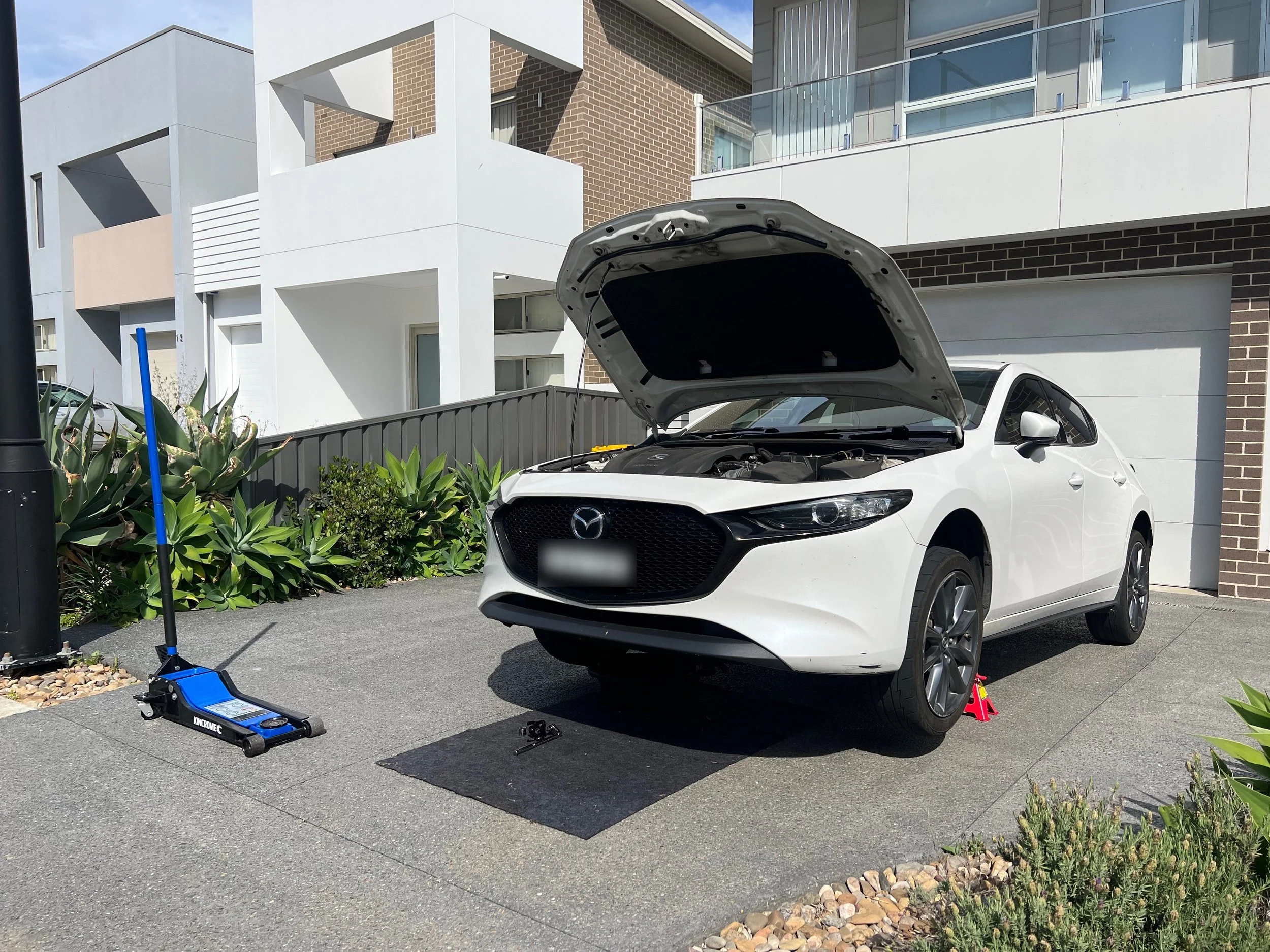 A white Mazda car with open hood parked on a driveway in front of modern residential buildings. A hydraulic jack and a car repair tool are placed near the front wheels, indicating the car is being serviced.