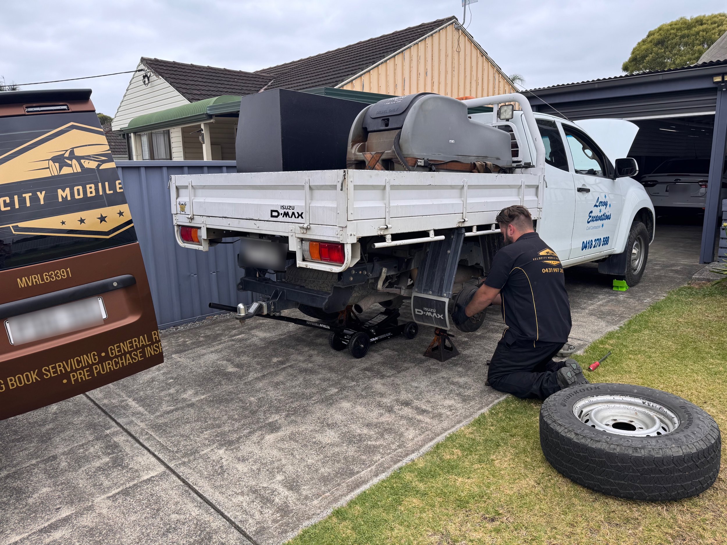 A mechanic repairing a white Isuzu D-Max pickup truck with the rear wheel removed, kneeling on the ground next to the vehicle, with a new tire and wheel nearby. The vehicle is parked on a driveway in front of a house, and another service vehicle is partially visible on the left.