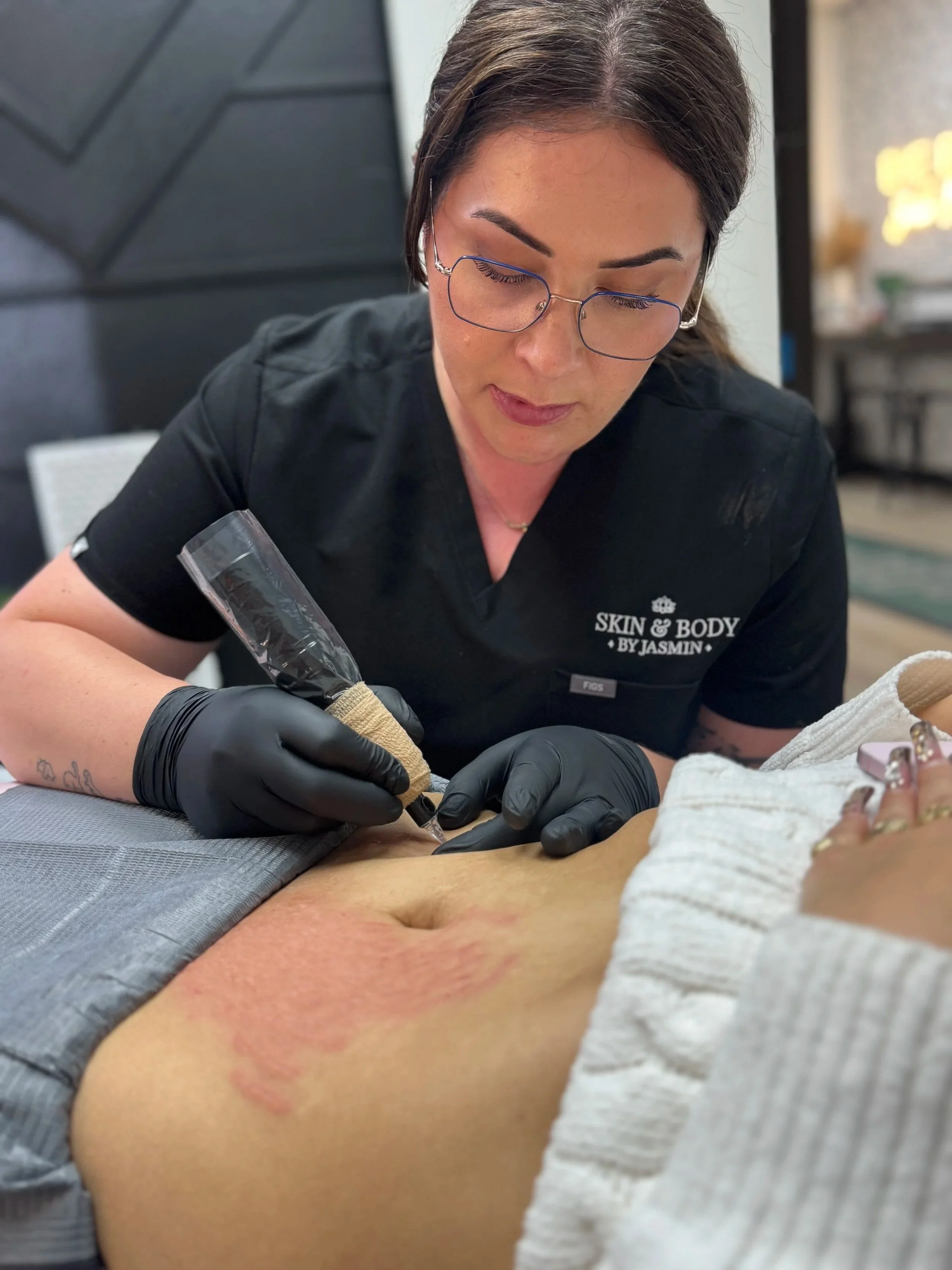 A woman with glasses and black gloves is tattooing a person's skin, which has red ink marks, in a professional tattoo studio.