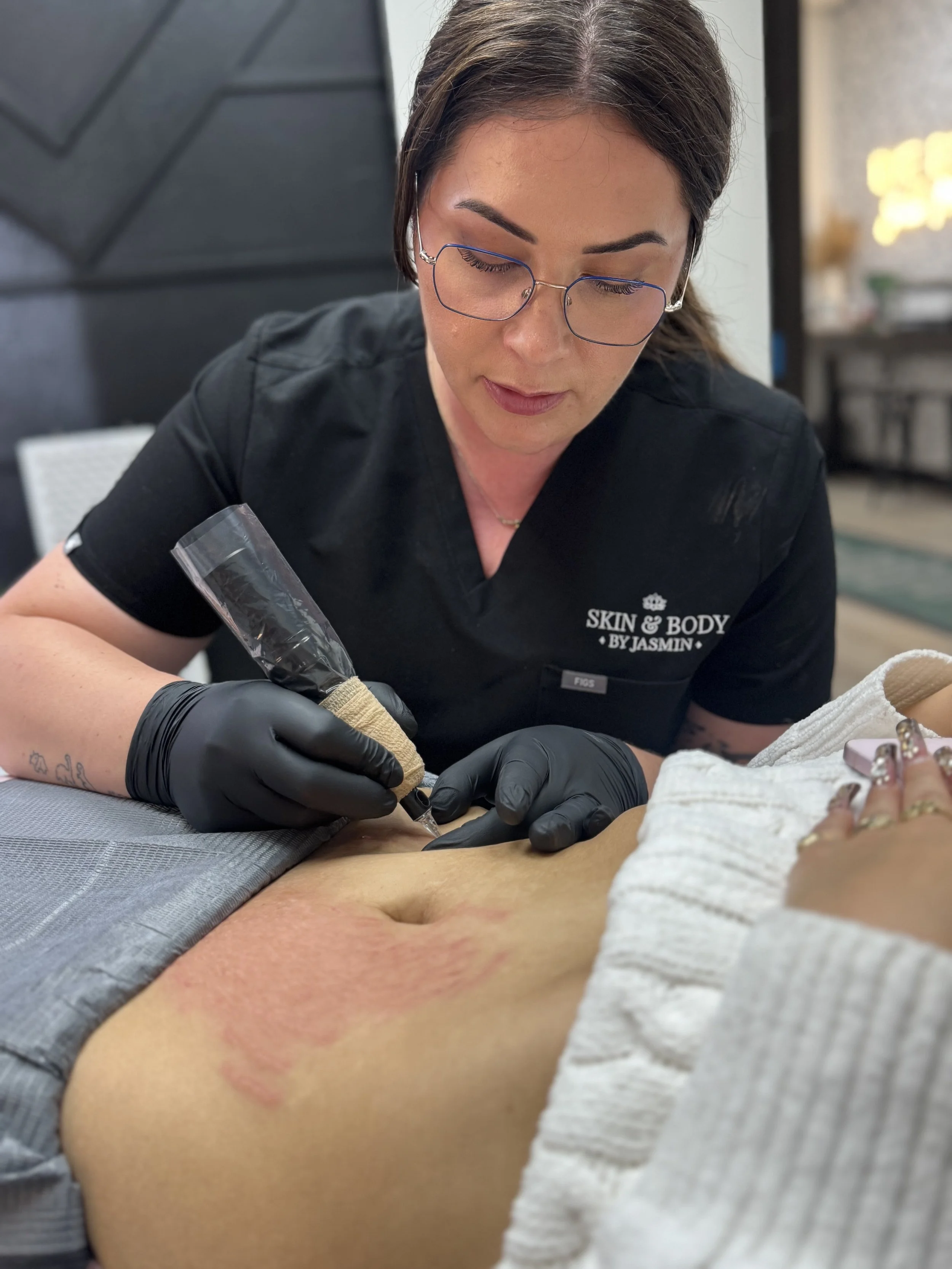 A woman with glasses and black gloves is tattooing a person's abdomen at a skincare and body tattoo studio.