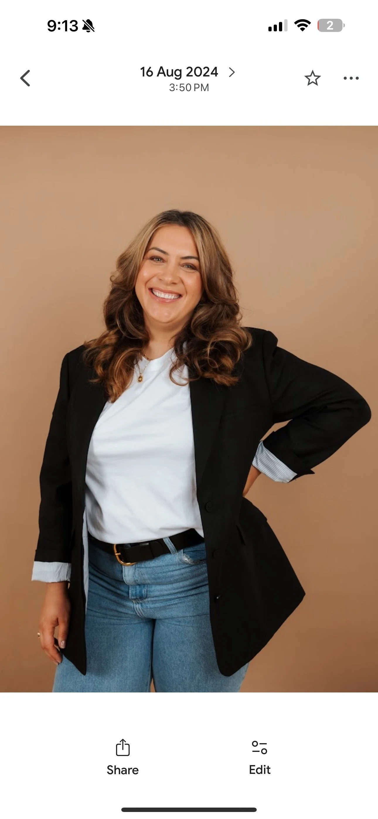 A woman with long wavy brown hair, wearing a white t-shirt, a black blazer with rolled-up sleeves, and blue jeans, standing against a beige background, smiling confidently with one hand on her hip.